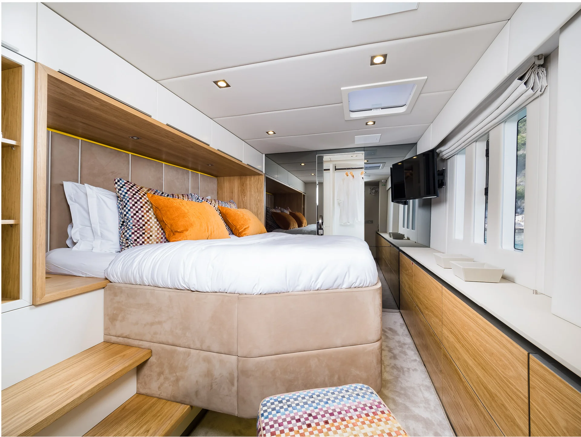 Modern cabin with a raised bed featuring orange and patterned pillows, wooden built-in storage, and a television mounted on the wall.