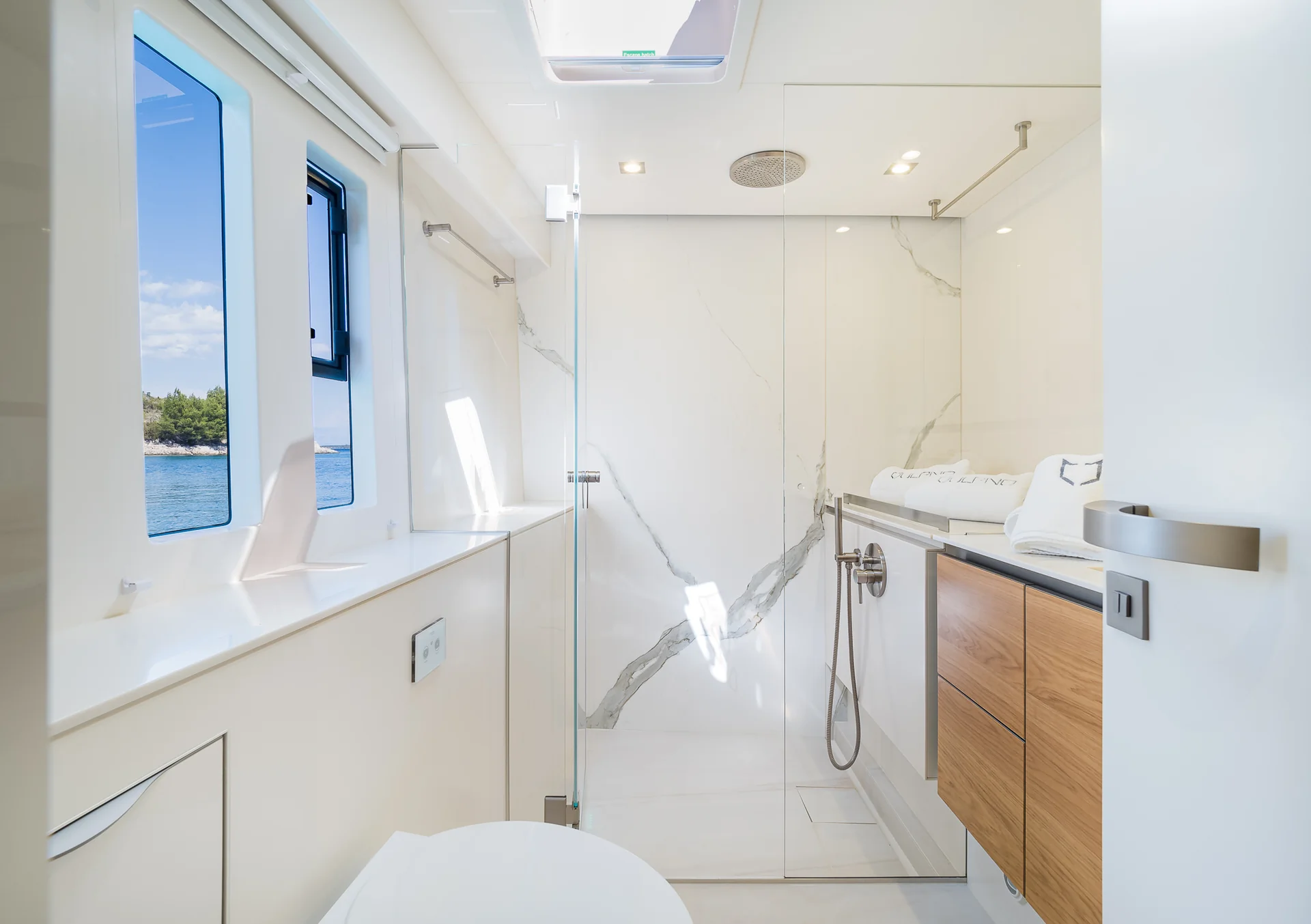 Modern yacht bathroom with a glass-enclosed shower, marble-patterned walls, and a view of the sea through a window.