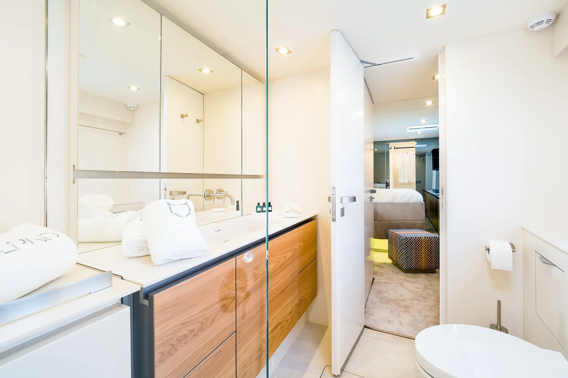 A modern bathroom with wood-grain vanity cabinets and a large mirror, showing a partial view of a bedroom through an open doorway.