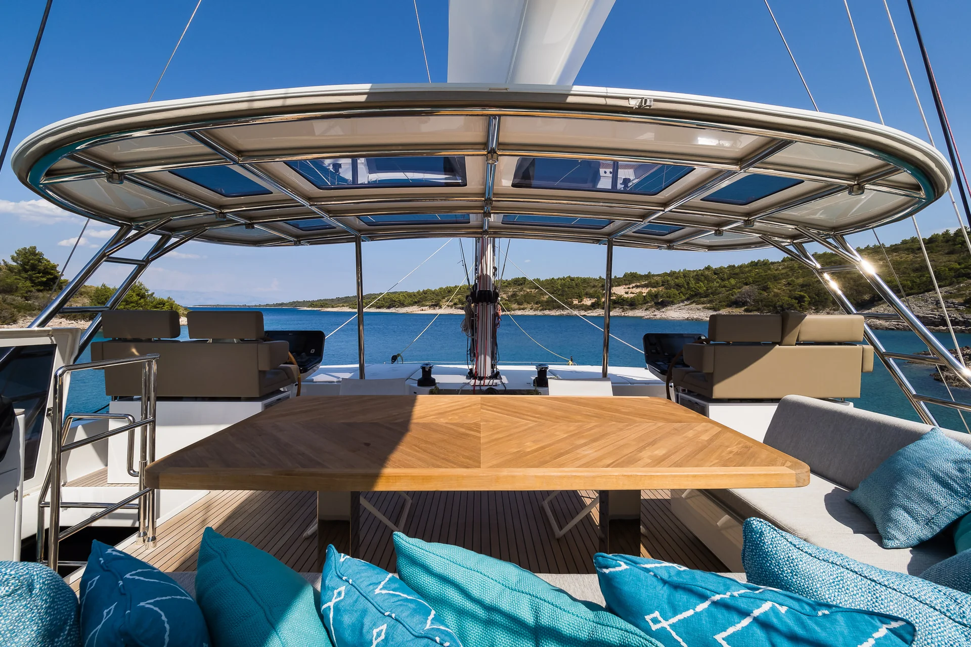 Outdoor seating area on a yacht deck with a wooden table, grey cushions, and patterned turquoise pillows, looking out over the blue sea.