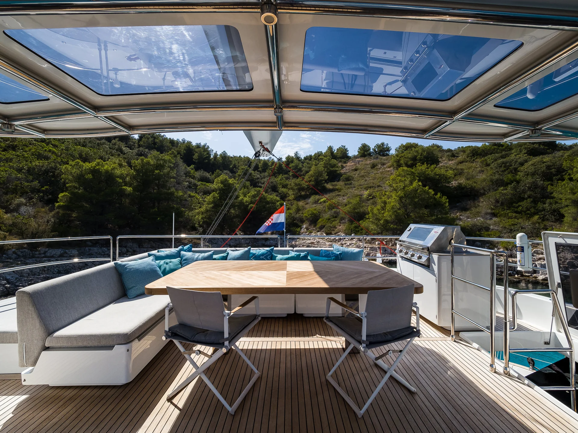 An outdoor dining area on a yacht deck featuring a wooden table, two chairs, a bench with blue cushions, and a stainless steel grill, with a view of a wooded coastline.