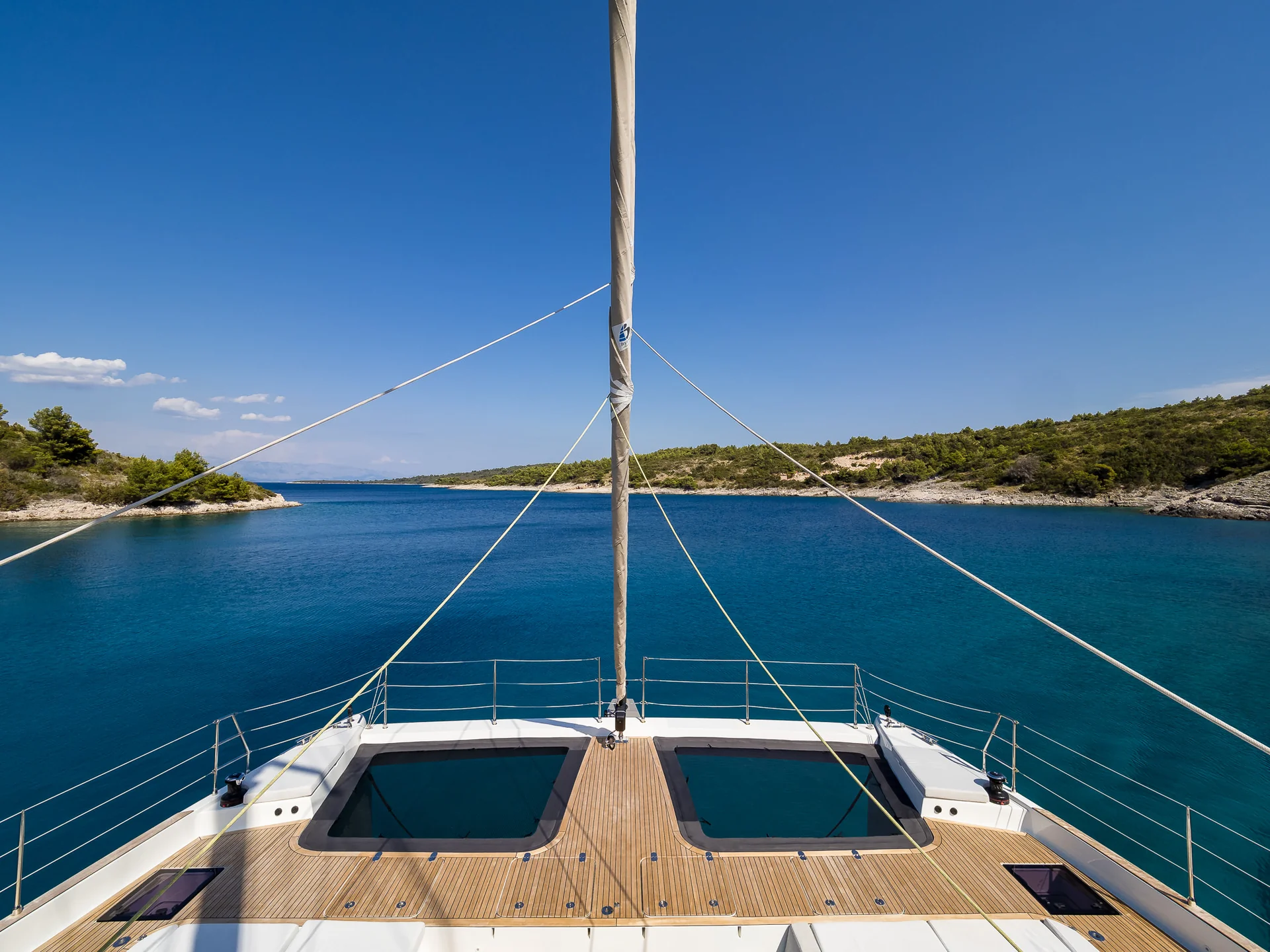 View from the bow of a catamaran sailing into a narrow, blue-water bay framed by rocky, tree-lined shores.