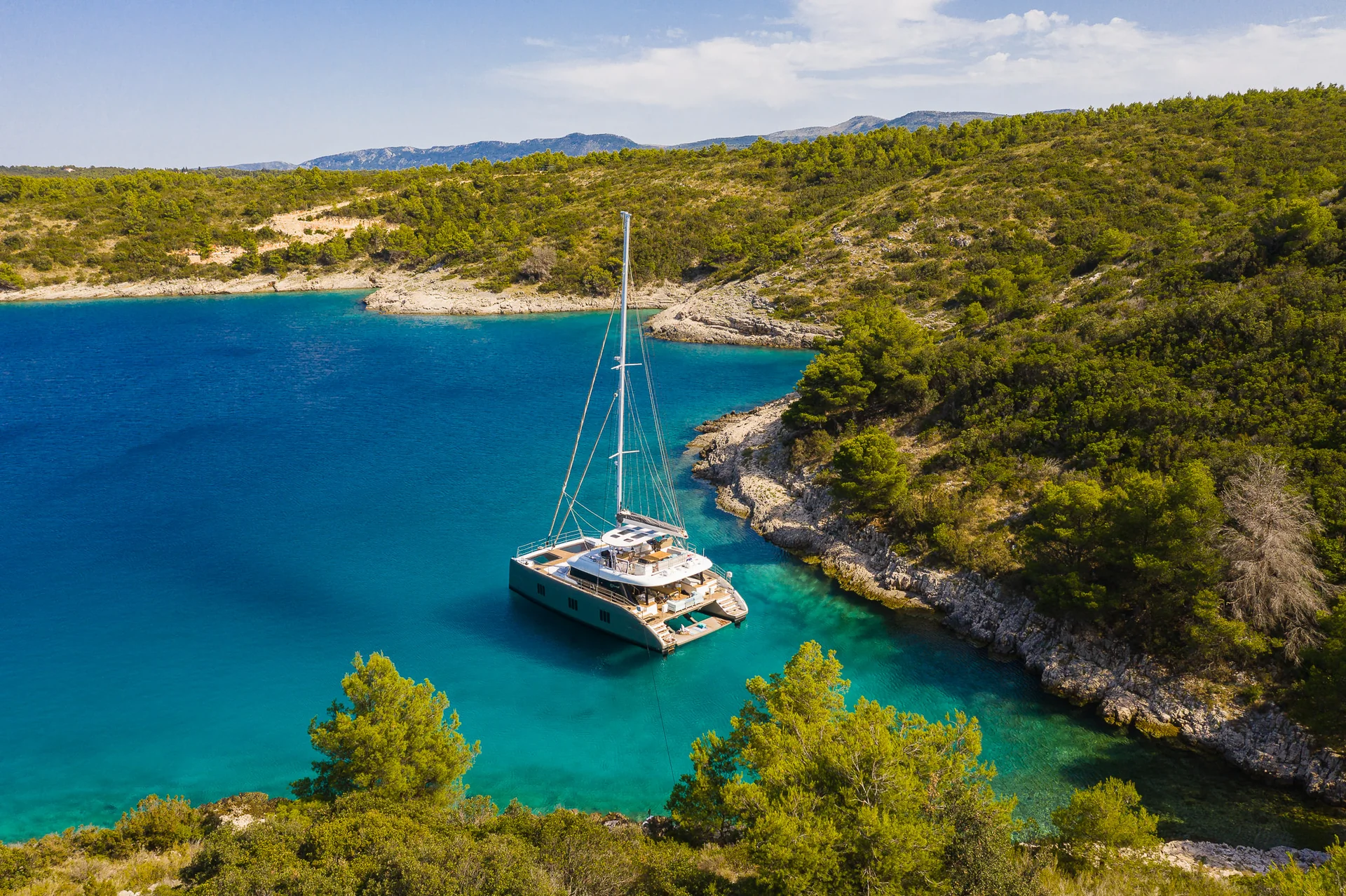 A catamaran anchored in a clear blue bay surrounded by a rocky, tree-lined coastline.