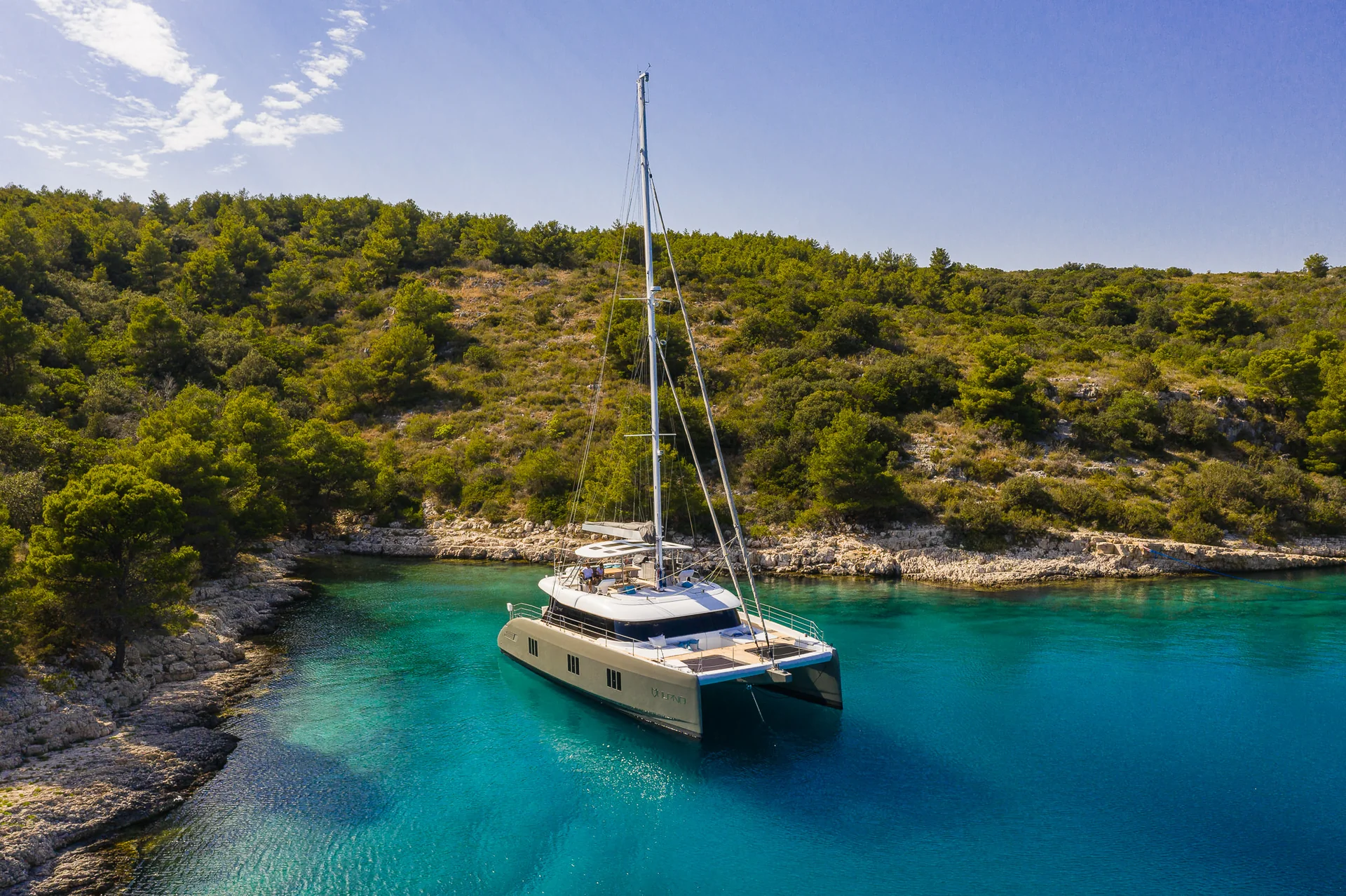 A catamaran sailboat anchored in a turquoise bay surrounded by a forested coastline.