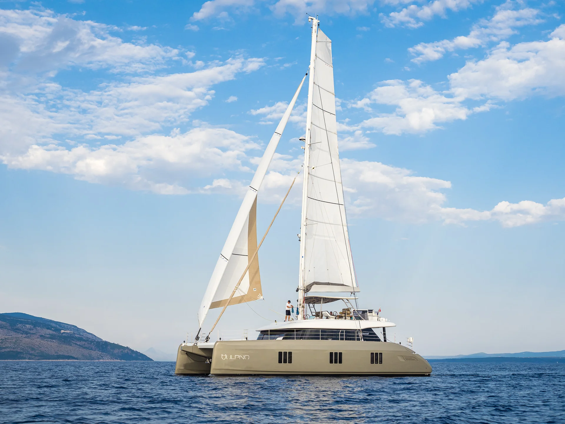 A tan sailing catamaran with a person on the upper deck moves across the open sea under a blue sky with scattered clouds.