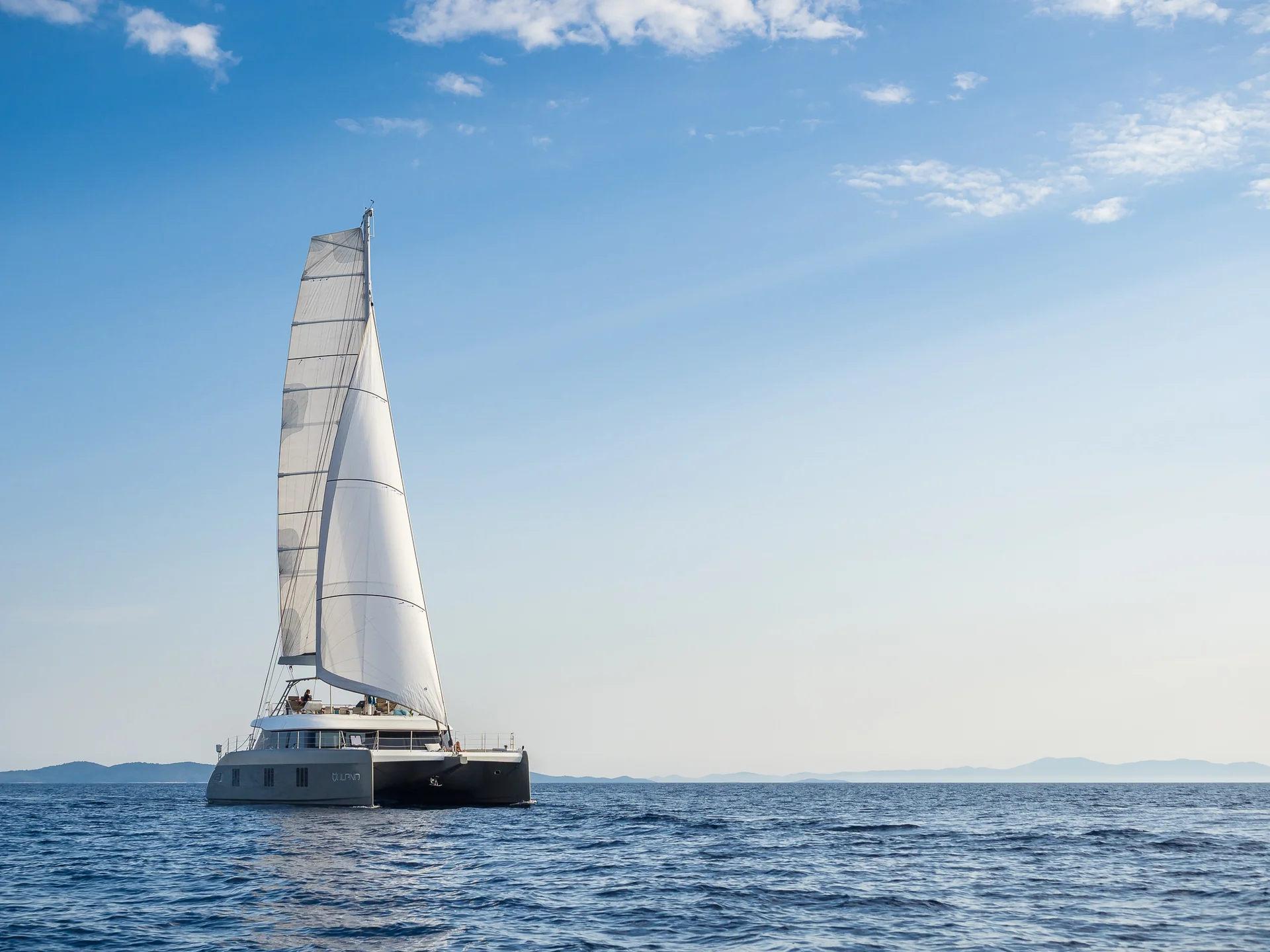 A large white catamaran sailboat cruising on the open sea under a blue sky.