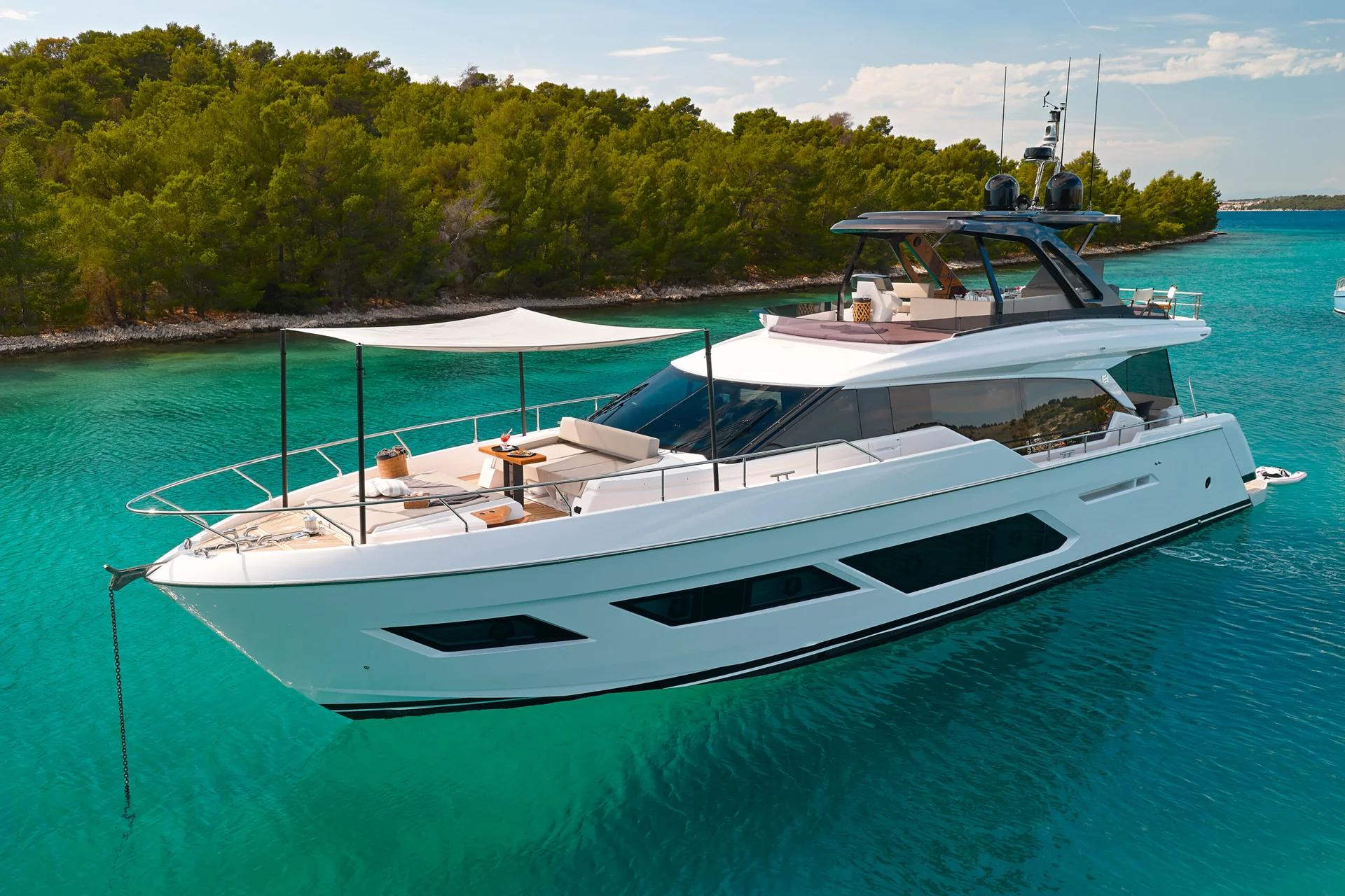 A white yacht anchored in clear turquoise water near a forested coastline.
