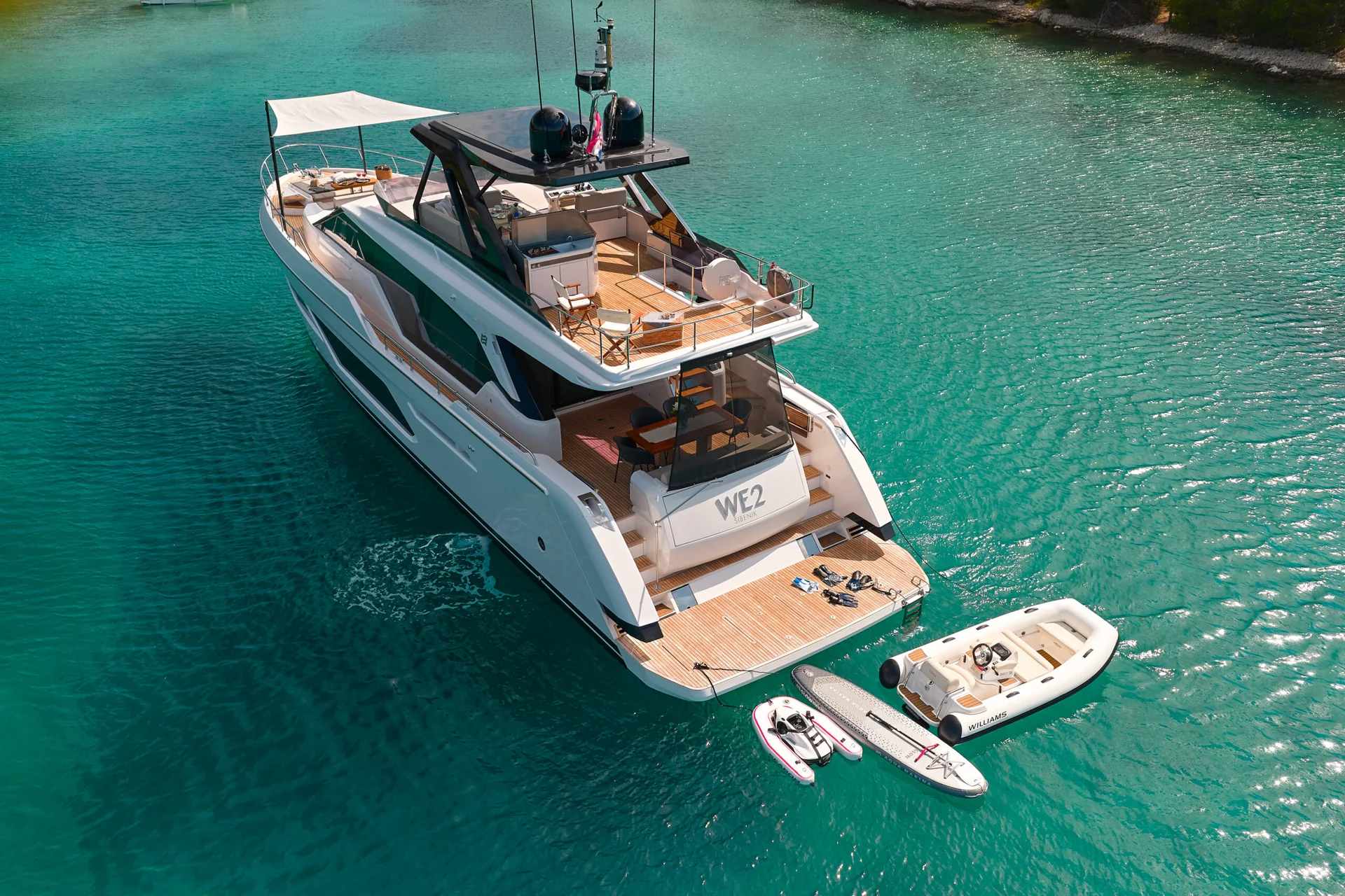 A white yacht named WE2 anchored in turquoise water, with a small tender, a paddleboard, and a towable tube floating nearby.