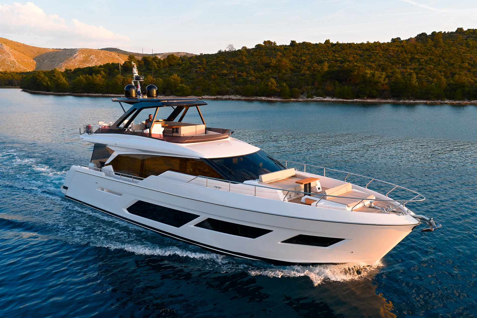 A white motor yacht cruising on calm water near a wooded shoreline.