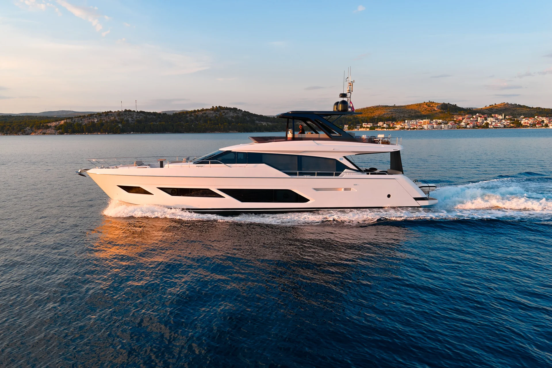 A white motor yacht cruising across the open sea near a coastline at sunset.