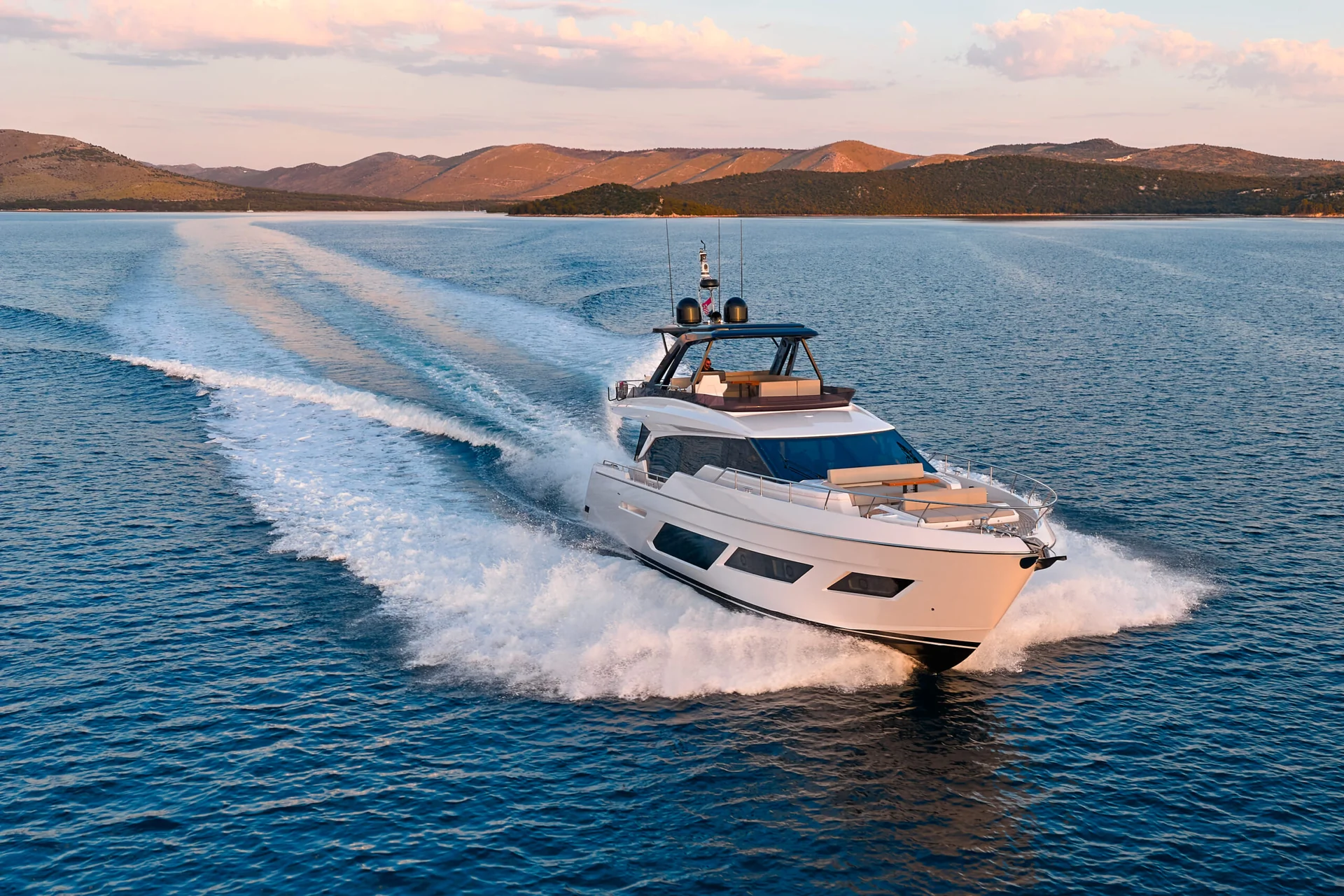 A white motor yacht cruising across the water, leaving a large wake behind it, with mountains in the background.