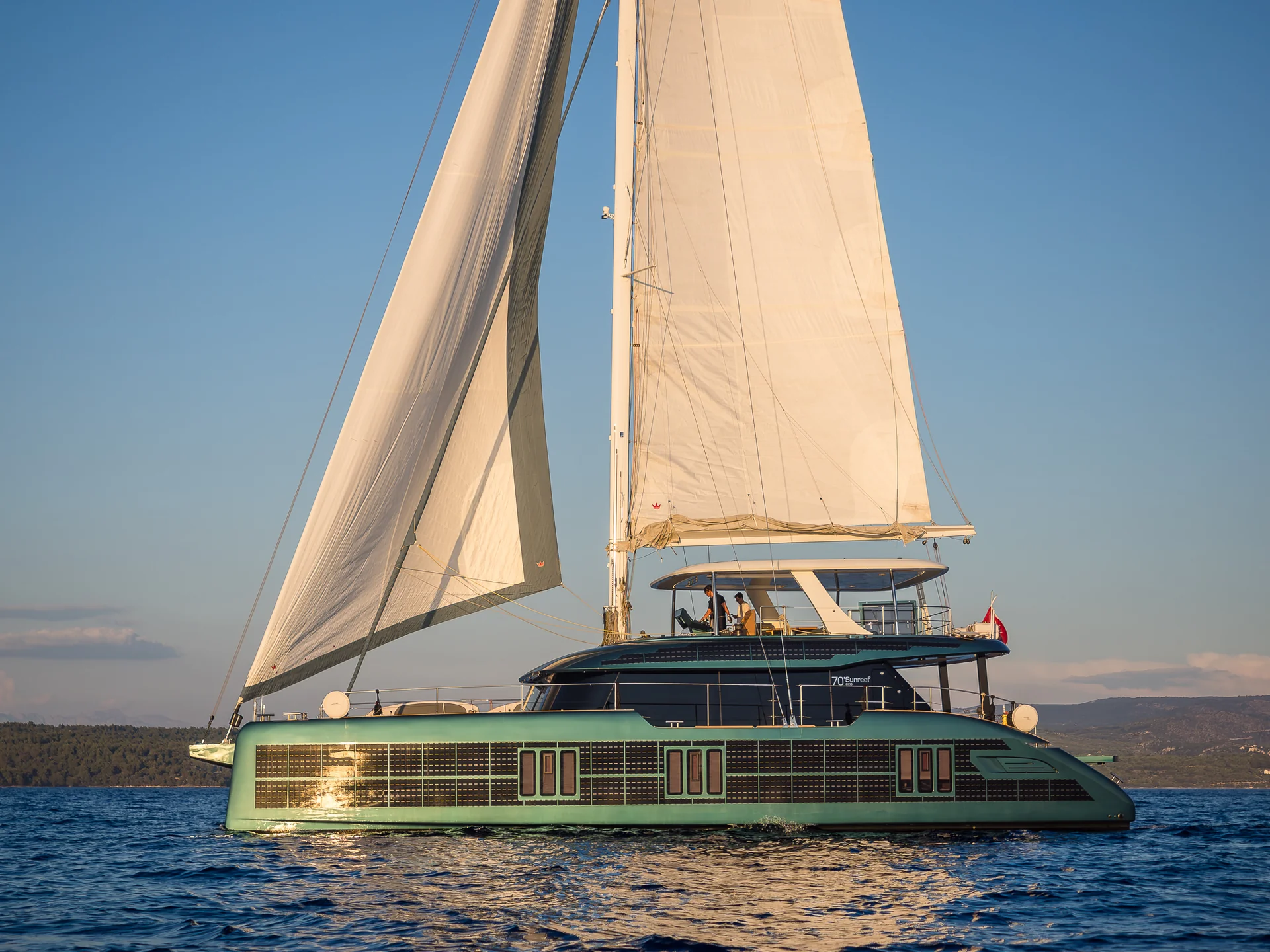 A teal catamaran sailboat with large solar panels integrated into its hull sailing on the water.