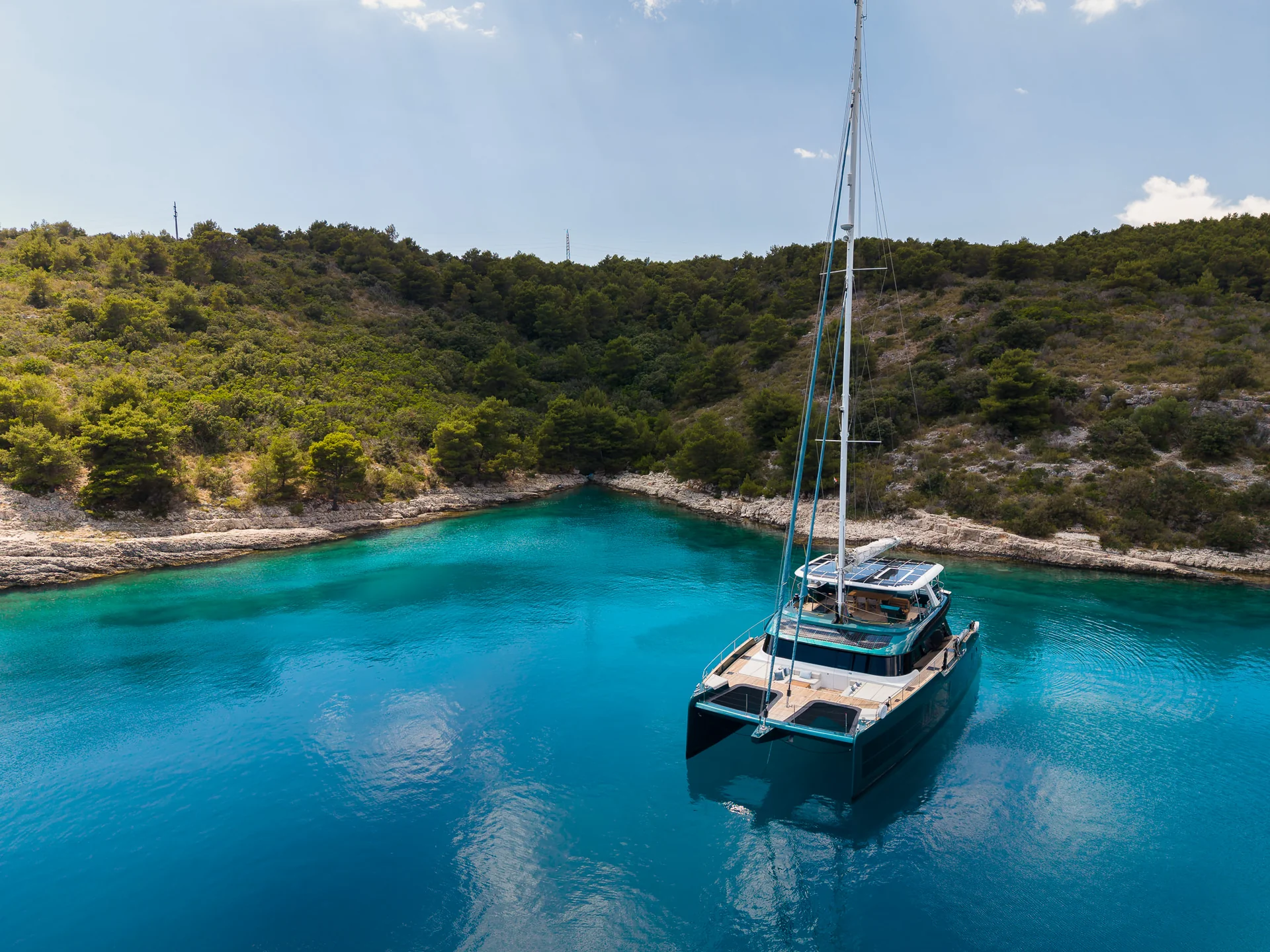 A large catamaran anchored in a clear, turquoise cove surrounded by a wooded coastline.