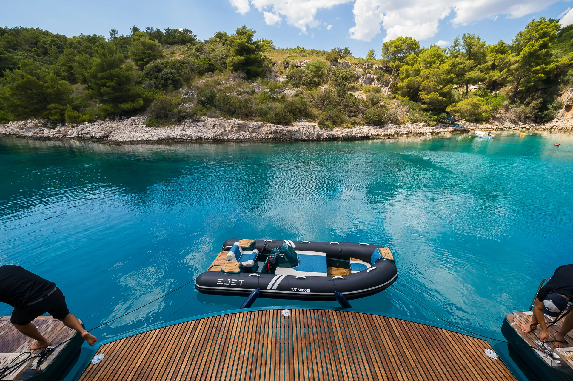 An inflatable jet tender tethered to the wooden swim platform of a boat, floating on clear turquoise water in a rocky cove.