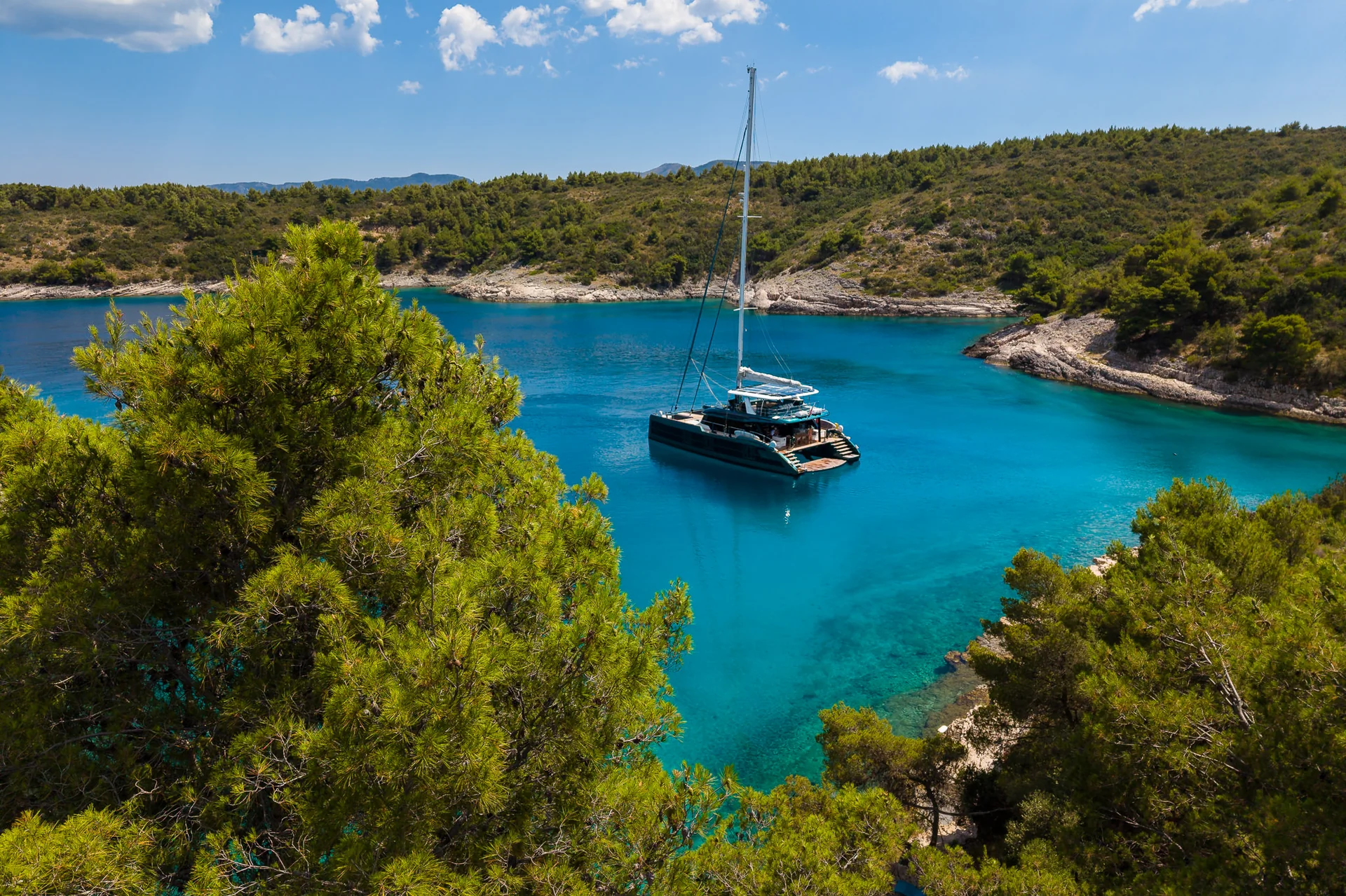 A catamaran anchored in a clear, turquoise bay surrounded by wooded hills.