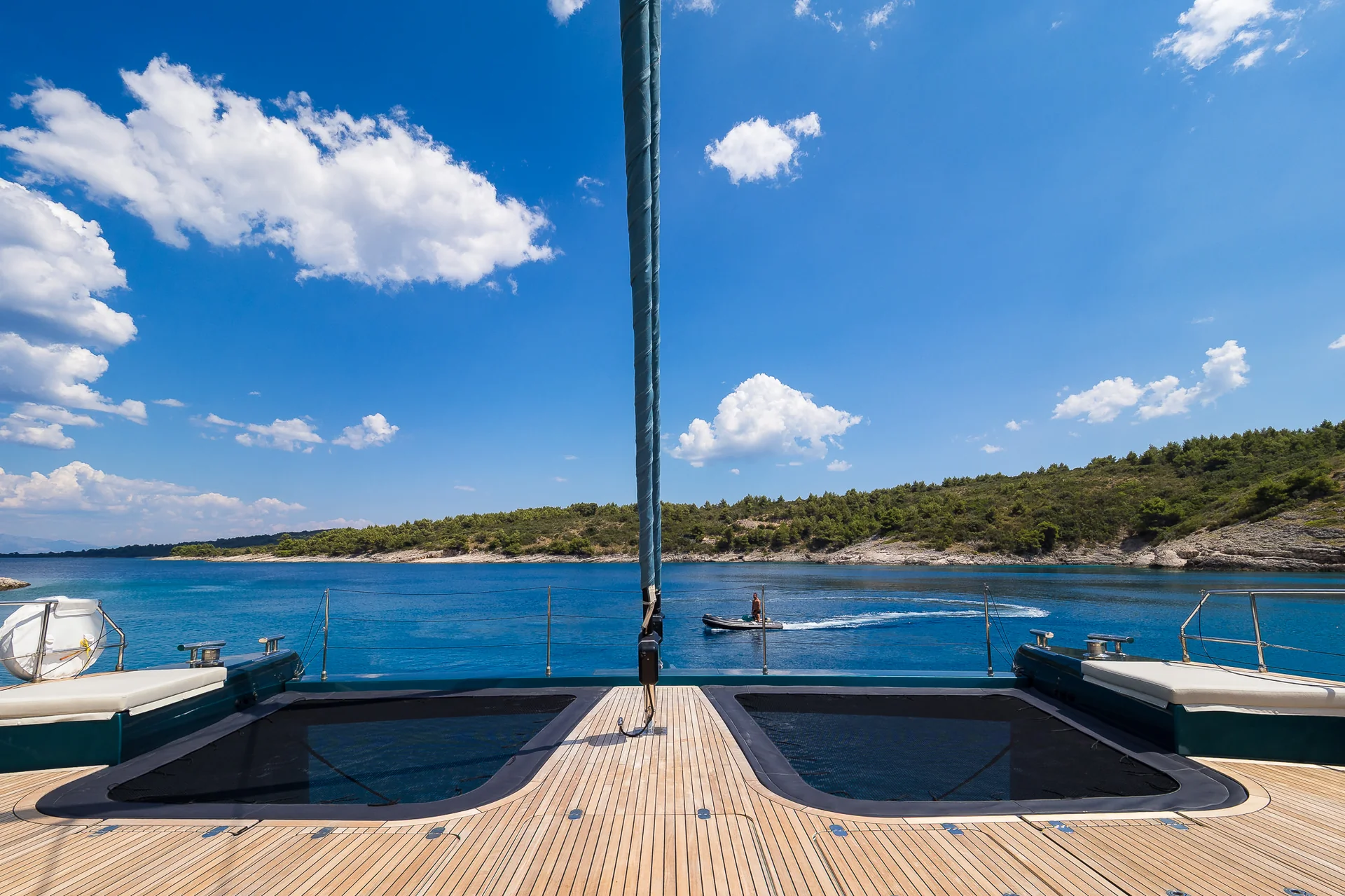 View from the deck of a catamaran looking out at a person riding a small motorboat across a blue bay towards a wooded coastline.
