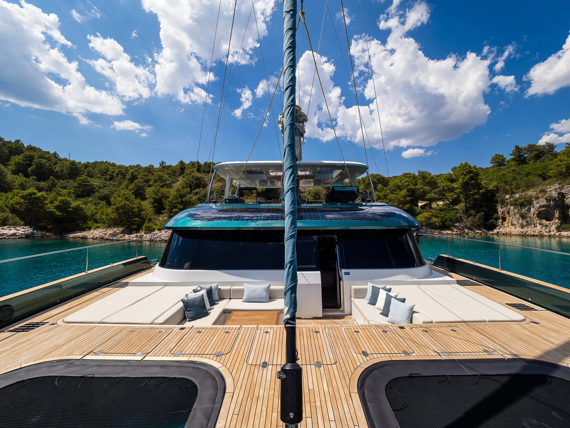 The deck of a catamaran featuring white lounge seating and a teak wood floor, facing a turquoise bay surrounded by trees.