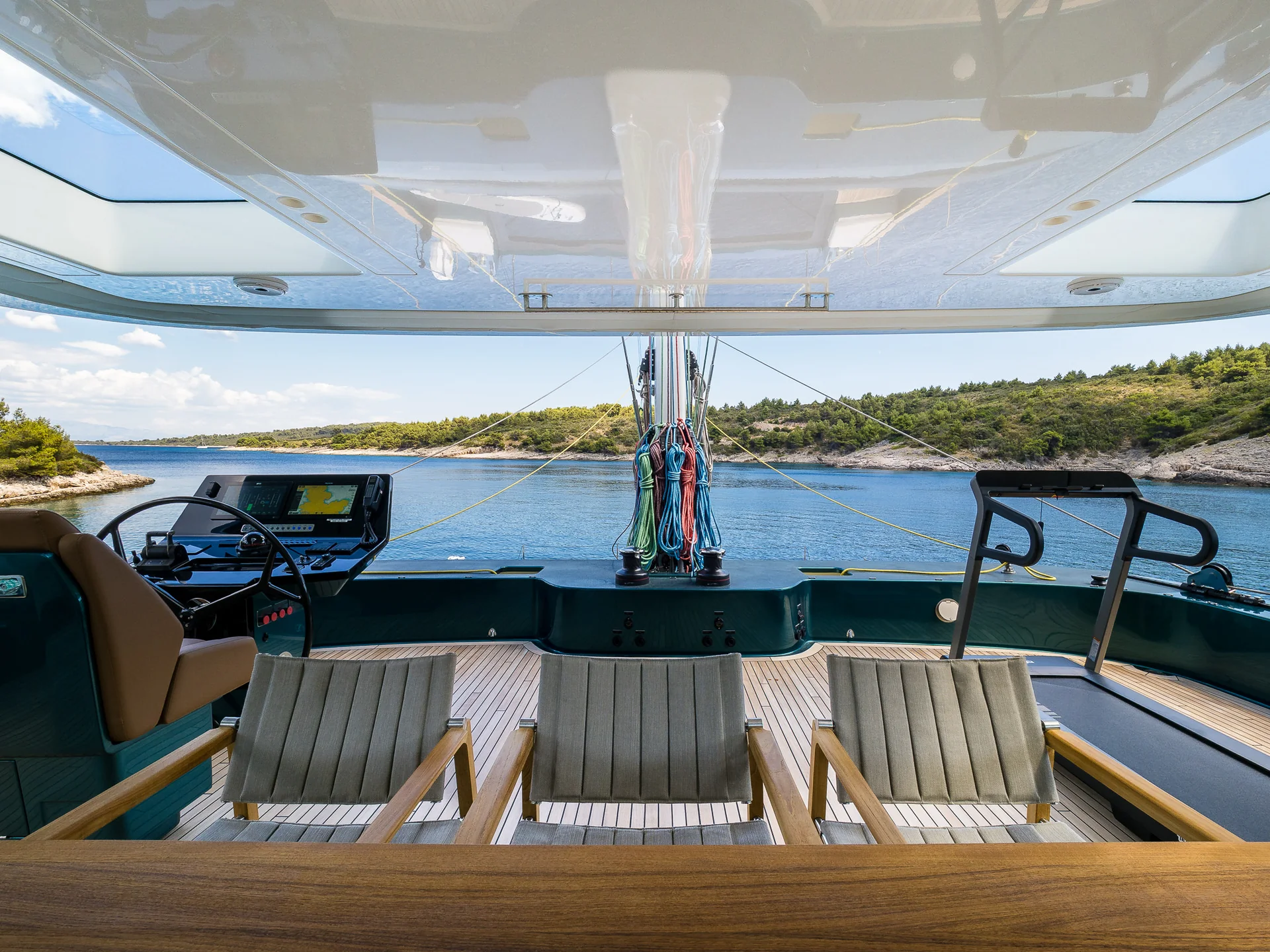 View from the shaded deck of a yacht looking out over a bay, featuring three chairs, a steering console, and a treadmill.