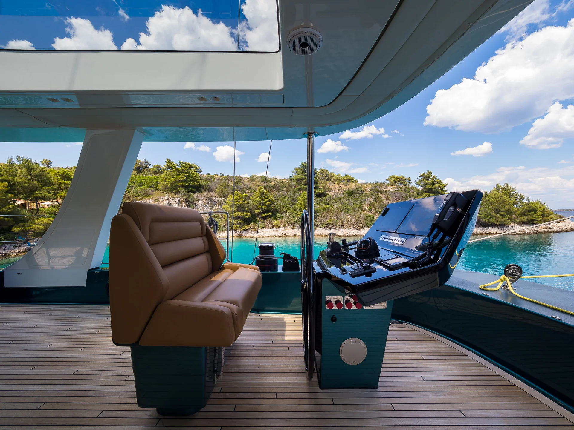 The helm station on a boat deck, featuring a tan leather captains chair, a navigation console with display screens, and a view of a coastal inlet with turquoise water and tree-covered hills.