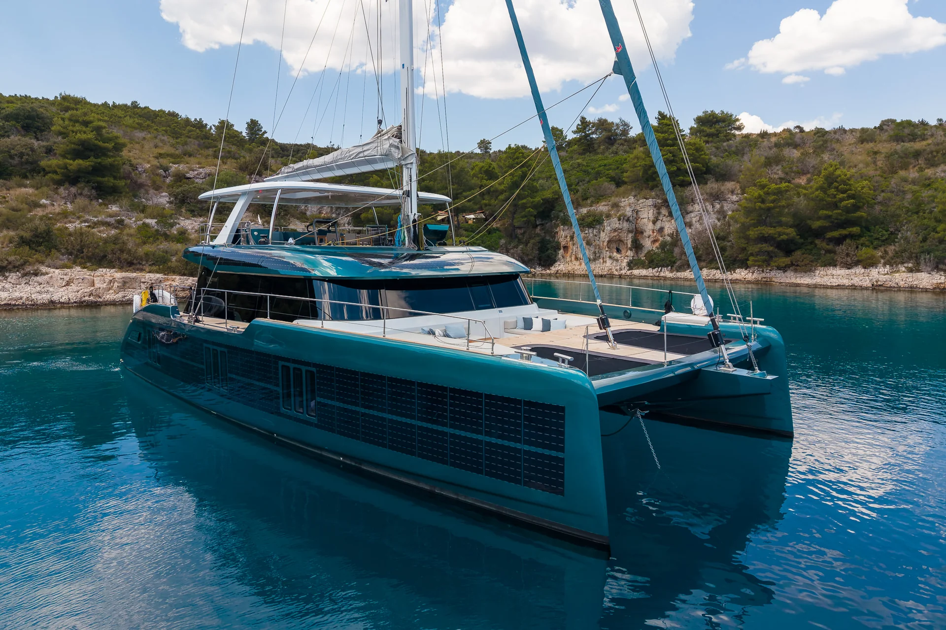 A large teal catamaran anchored in a calm cove surrounded by rocky, tree-covered hills.