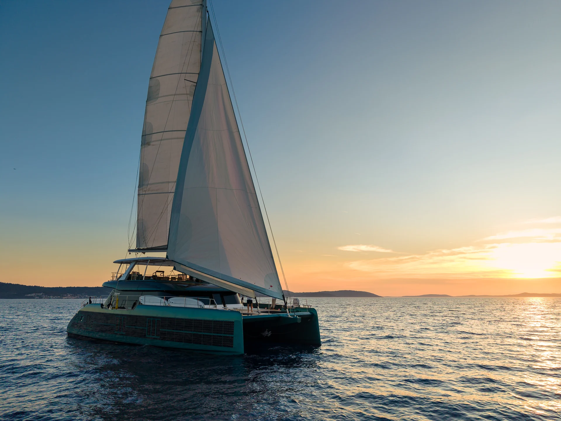 A sailing catamaran under full sail on the ocean at sunset.