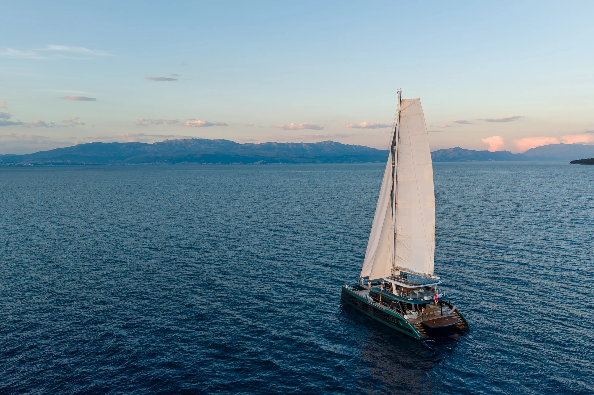 A sailing catamaran cruising on open water with distant mountains in the background at sunset.