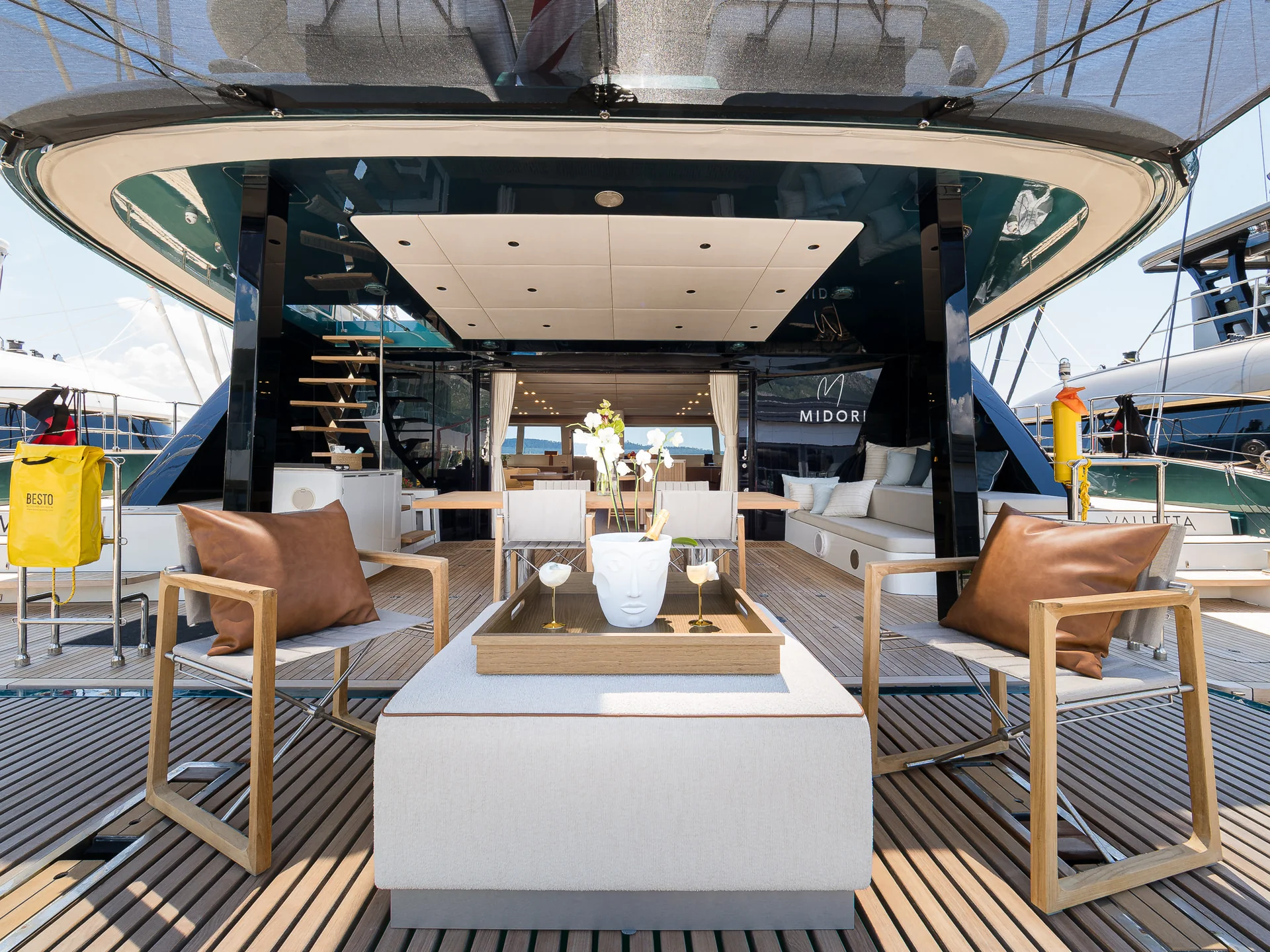 The rear deck of a yacht featuring two armchairs with brown cushions, a low white ottoman, a wooden tray with a decorative vase, and a dining table with chairs.