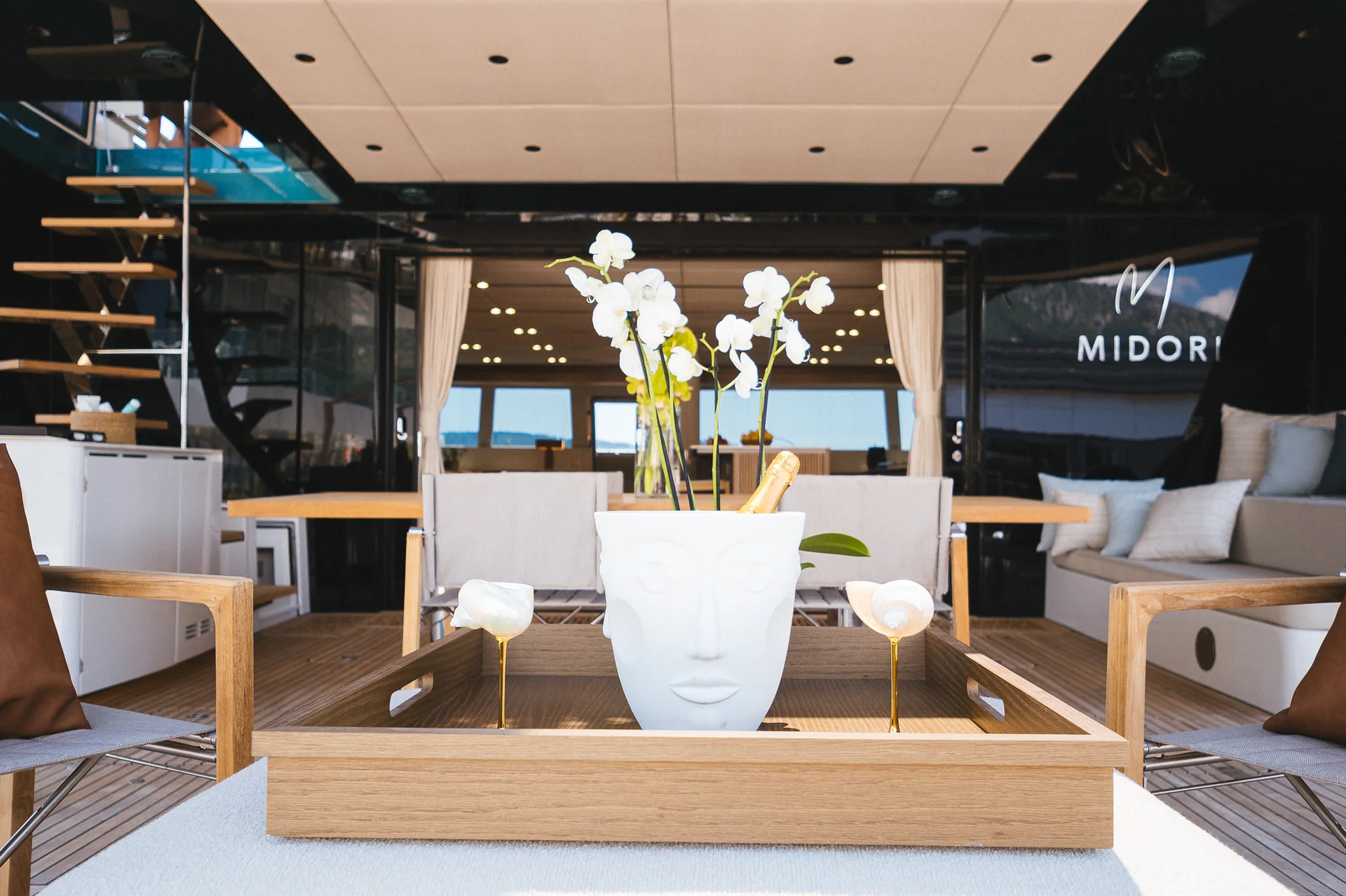 A white face-shaped planter containing white orchids and a champagne bottle sits on a wooden tray on the deck of a yacht.