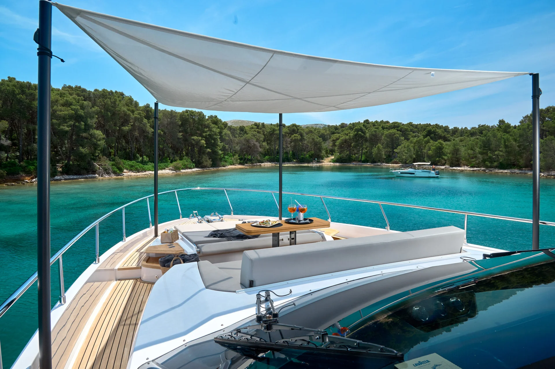 The deck of a yacht featuring a table with drinks and snacks, shaded by a canopy, overlooking a clear turquoise bay surrounded by pine trees.