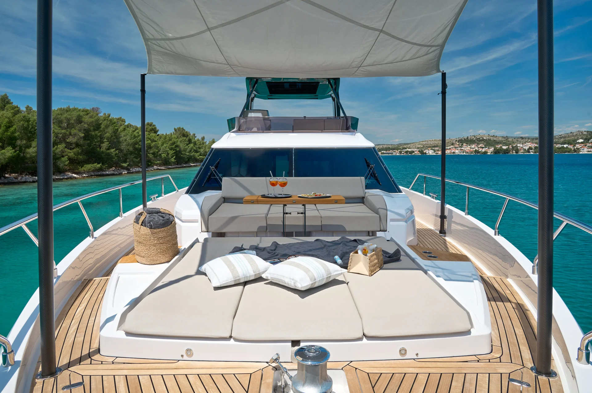 View from the deck of a yacht showing a shaded lounge area with sun pads, seating, and a small table with drinks, overlooking turquoise water and a coastline.