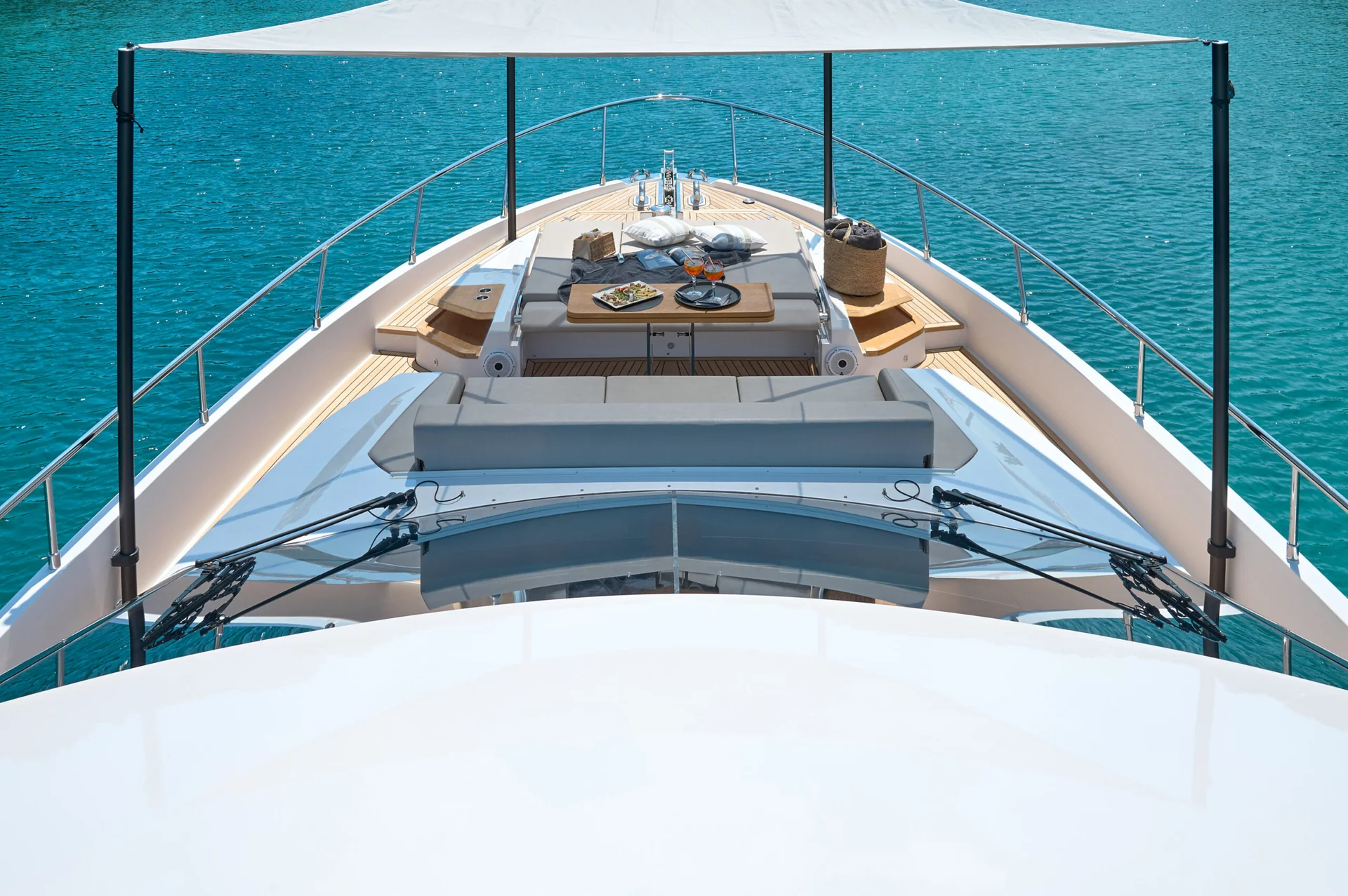 View from the top deck of a yacht looking toward the bow, which features cushioned seating, a table with food and drinks, and a sun shade over turquoise water.