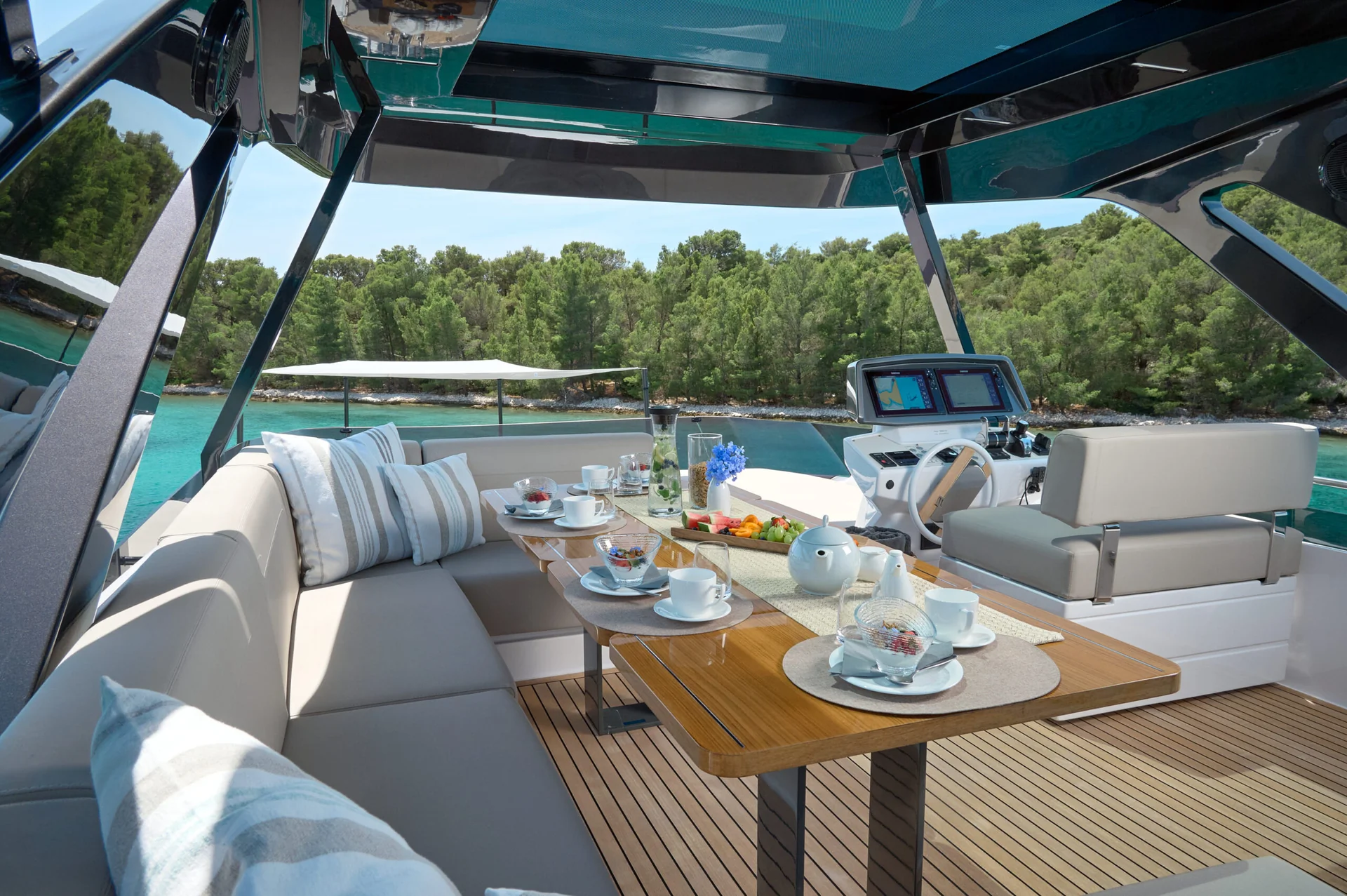 An outdoor dining table set for breakfast on the deck of a yacht, overlooking a turquoise bay and a pine forest.