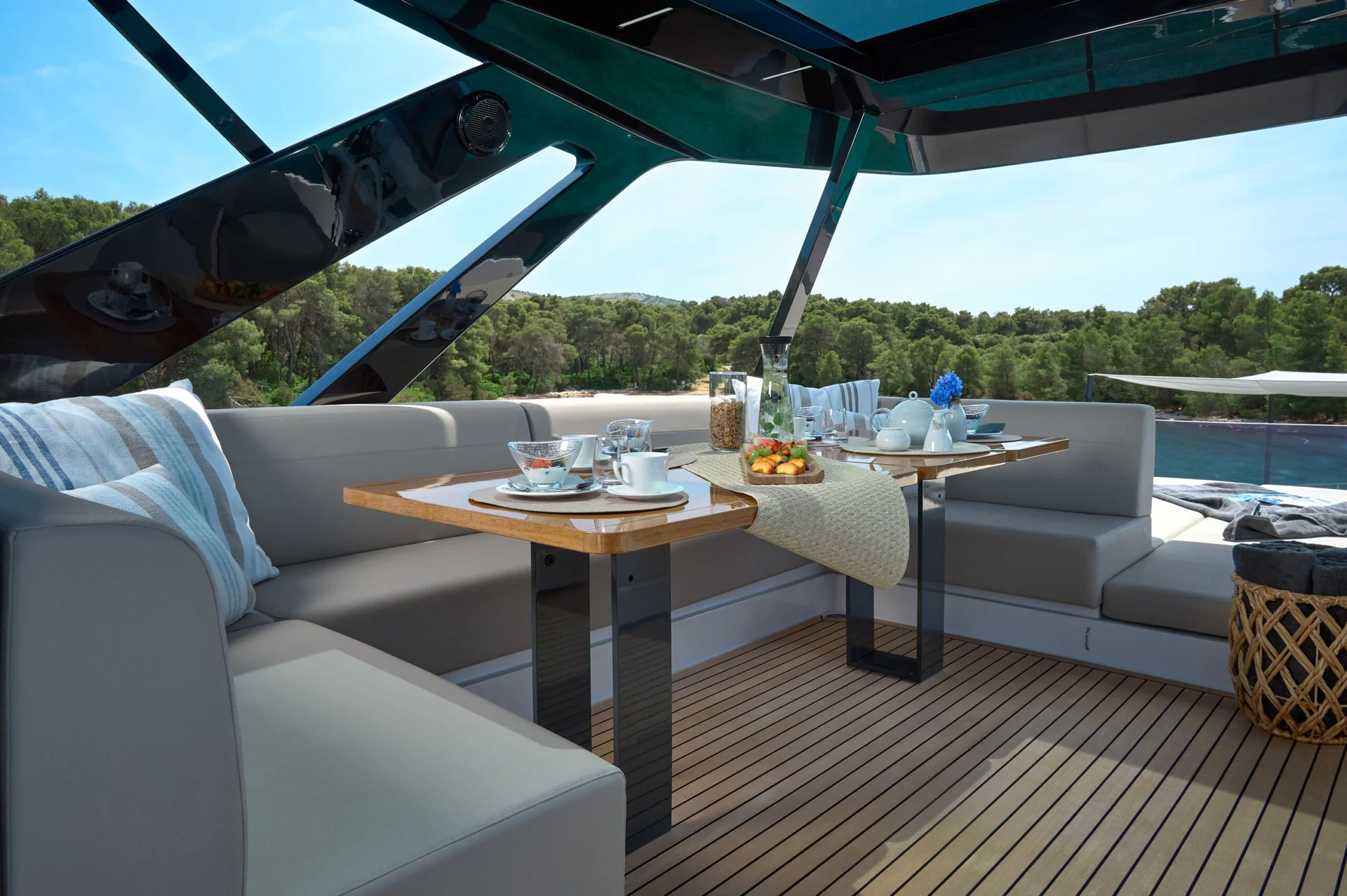 Dining area on a boat deck with gray cushioned seating, a wooden table set for tea, and a view of trees and water.
