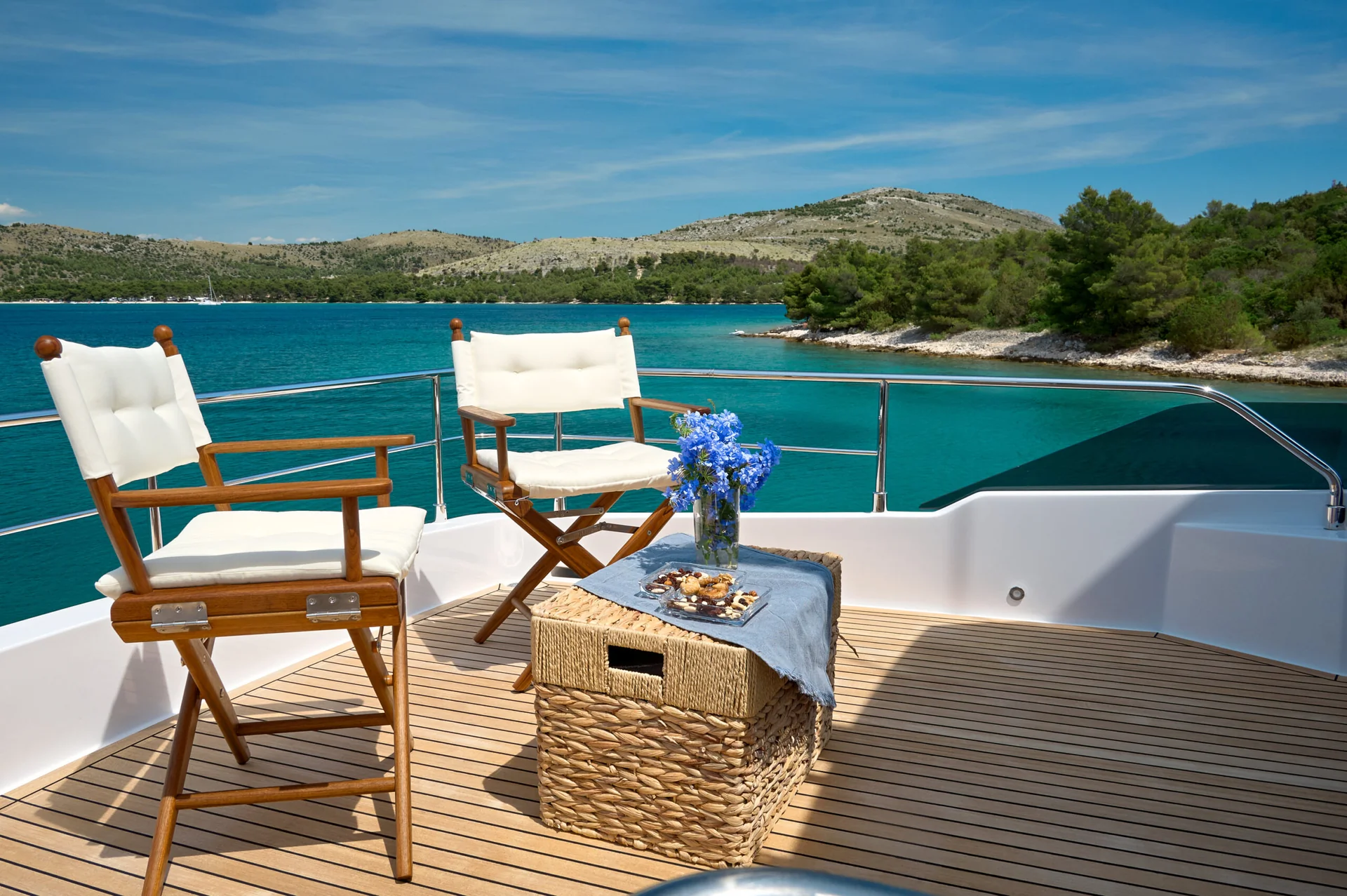 Two wooden folding chairs, a wicker storage ottoman with a blue cloth, a vase of blue flowers, and a small tray of snacks on the deck of a yacht overlooking a bay.