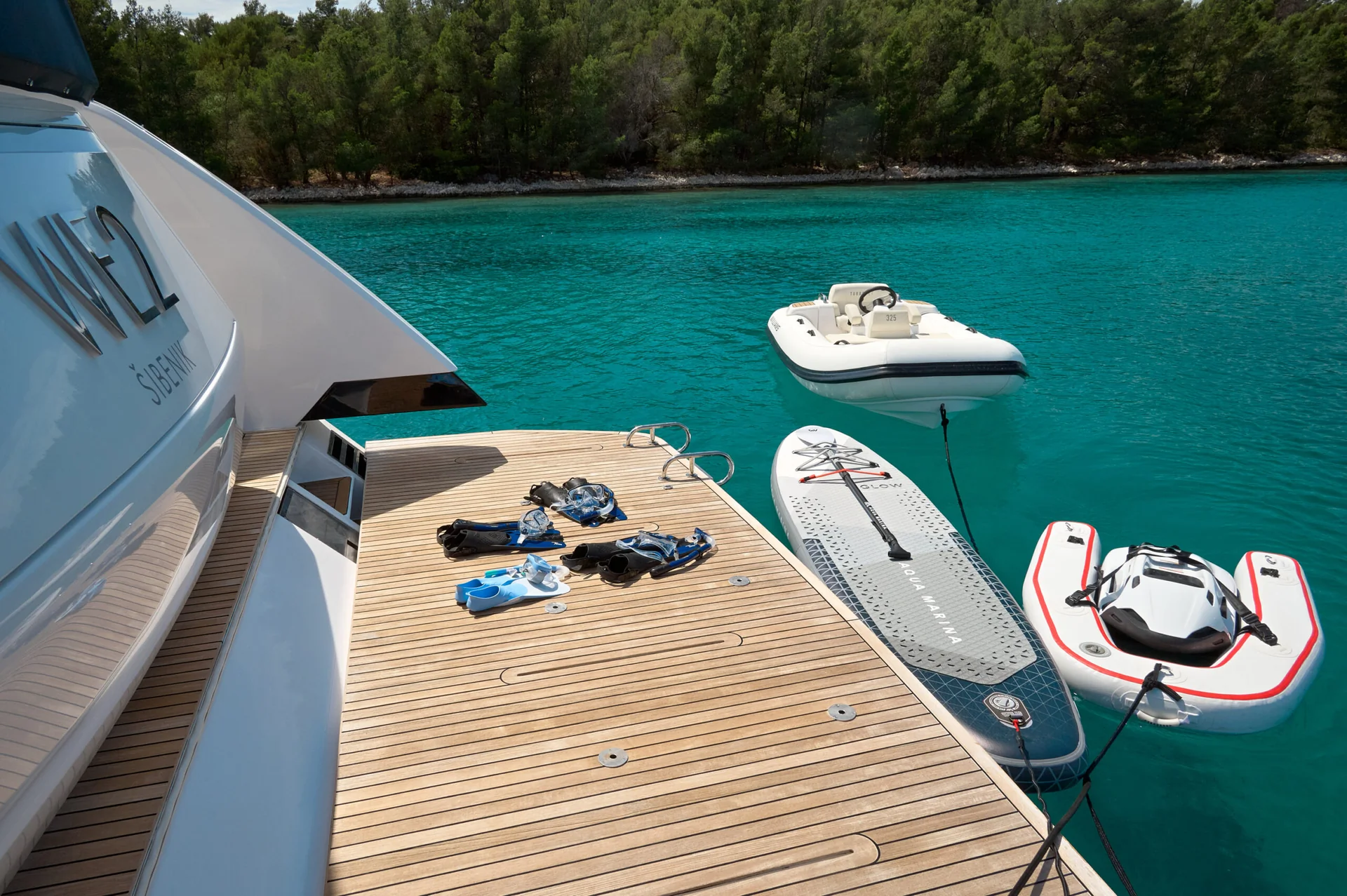 Teak deck of a yacht with snorkeling gear, a paddleboard, and two small inflatable boats floating in turquoise water.