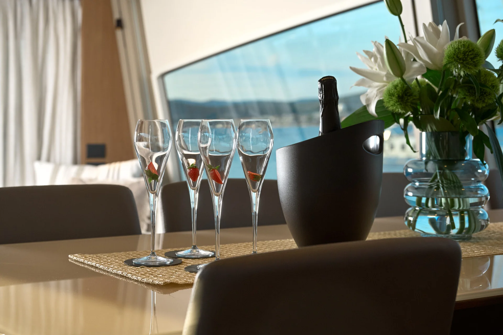 Four empty champagne flutes each containing a single strawberry slice, next to a black champagne bucket and a vase of flowers on a table.