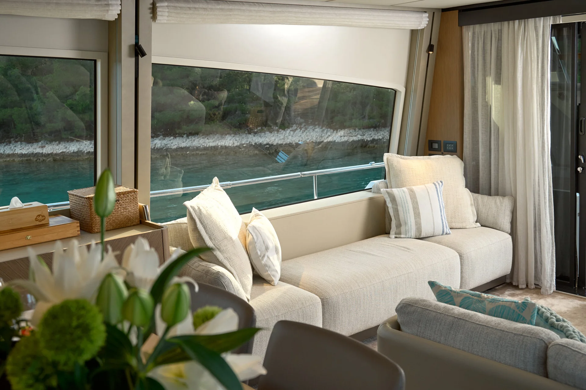 A lounge area inside a boat with a long beige sofa, white cushions, and large windows overlooking the water.