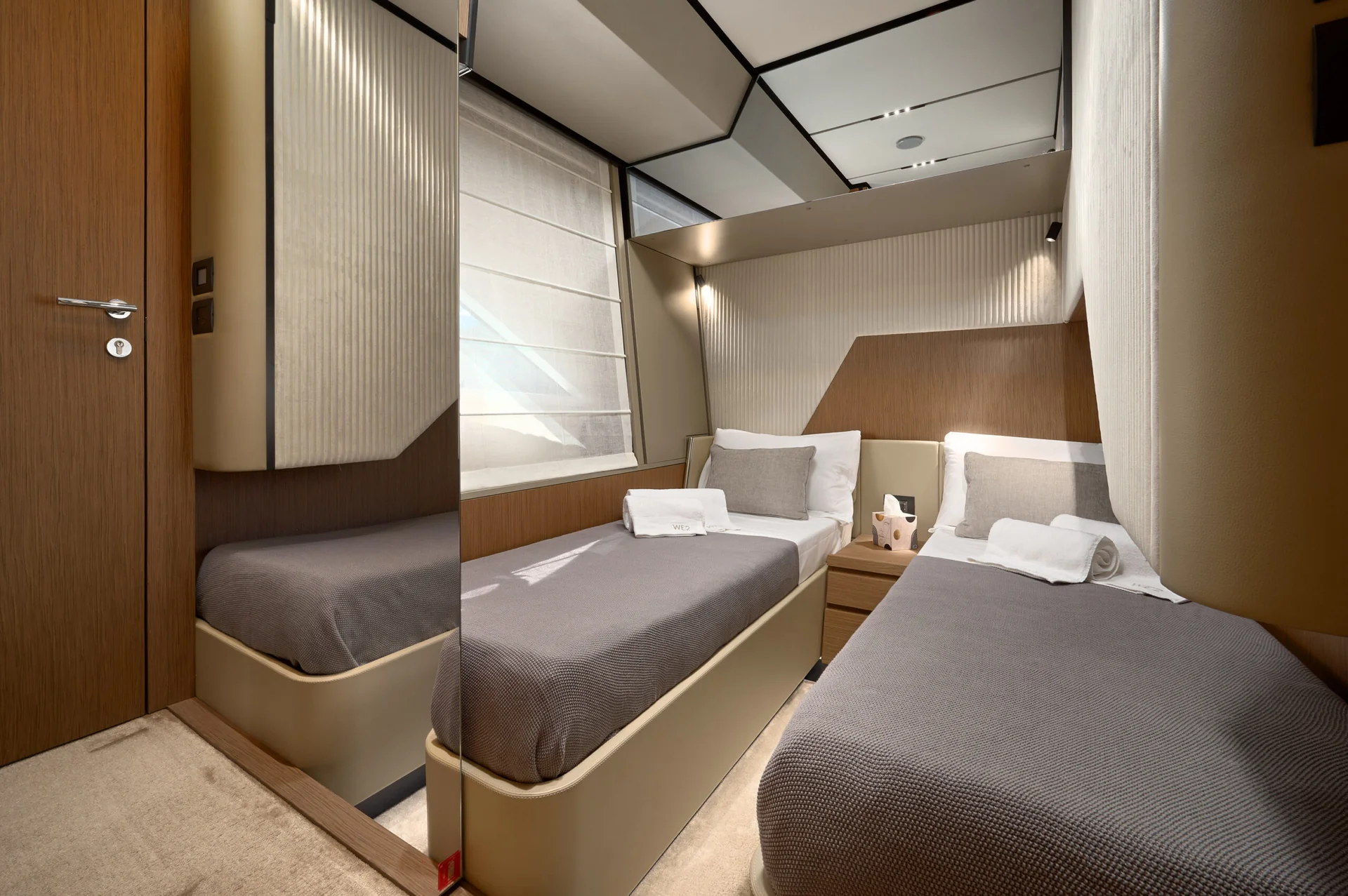 A cabin on a yacht featuring two single beds with grey bedspreads and white towels, separated by a nightstand.