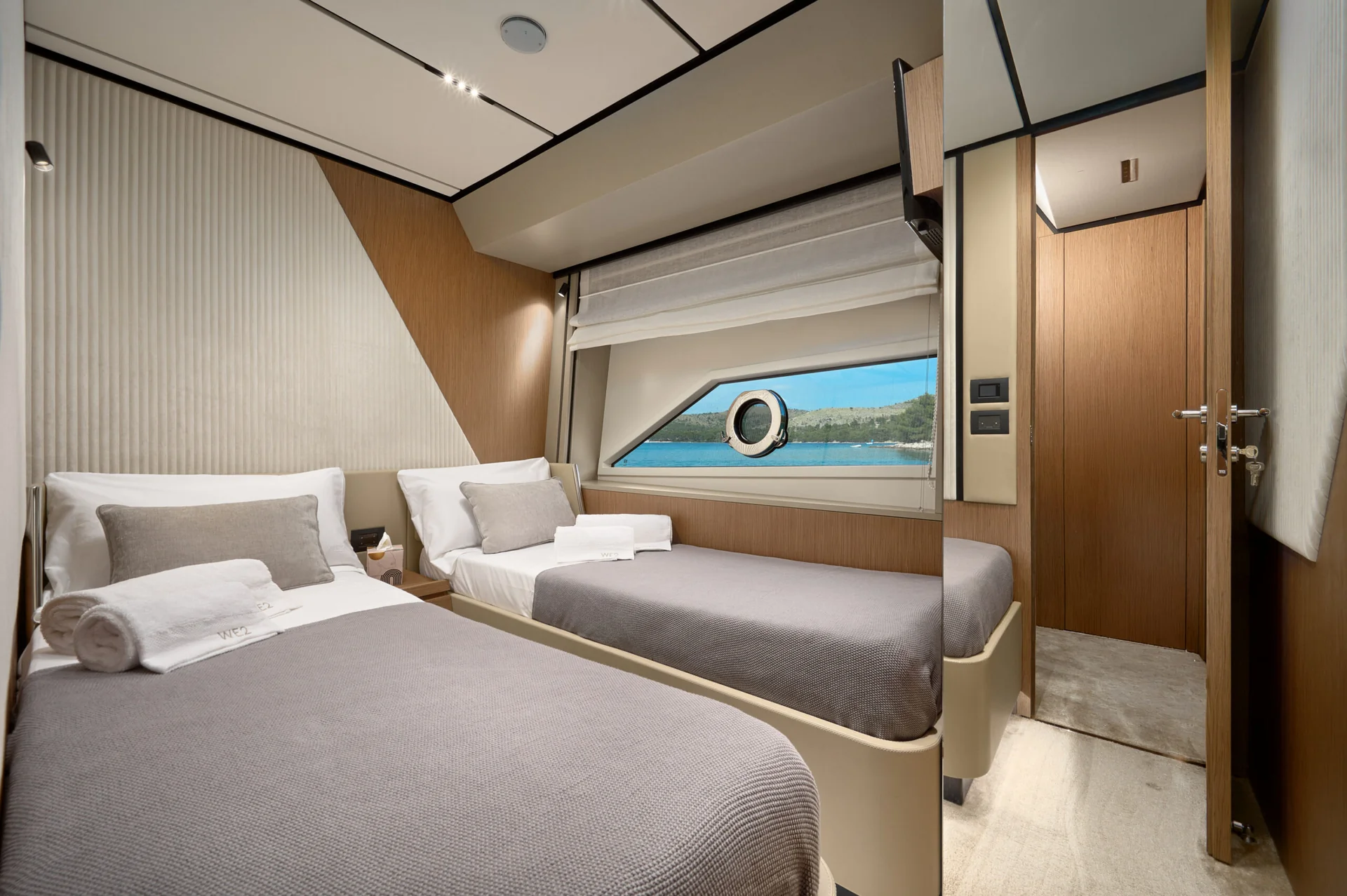 Interior of a yacht cabin featuring two twin beds with gray bedding and a view of the ocean through a porthole window.