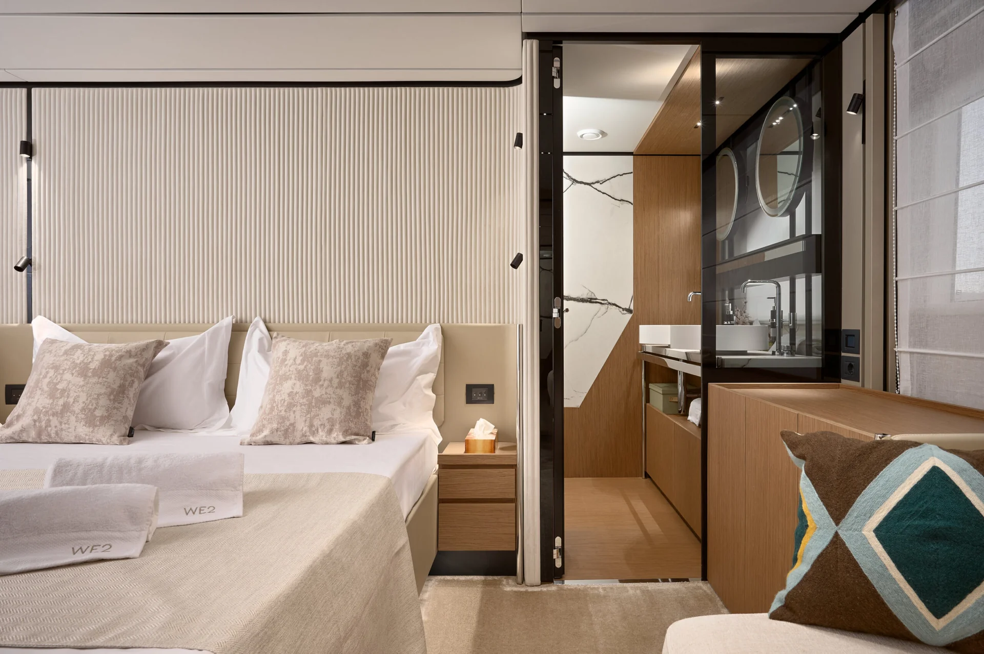 A bedroom featuring a bed with patterned pillows and folded white towels, with a sliding door revealing an adjacent bathroom with a marble-patterned wall and a vessel sink.