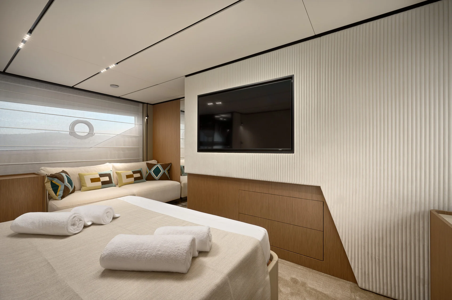 A modern bedroom on a yacht with a flat-screen TV mounted on a textured wall, a bed with folded white towels, and a sofa with patterned throw pillows.
