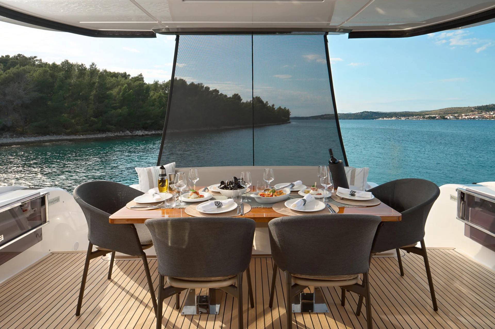 Dining table set for four on the deck of a yacht, overlooking a calm bay with wooded shores.