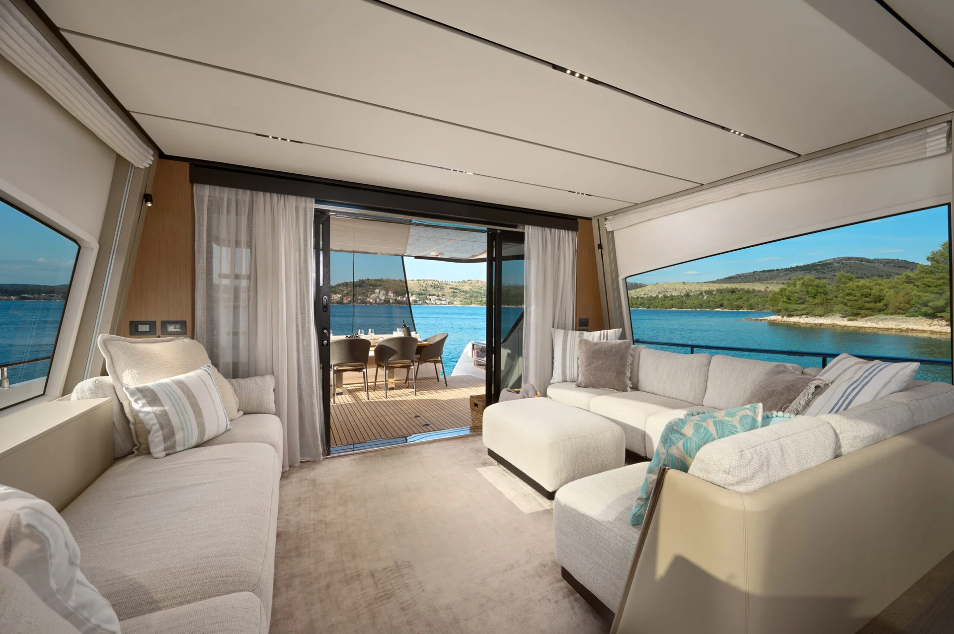 The interior lounge of a yacht with plush white seating areas facing an open deck and a view of the ocean and coastal hills.