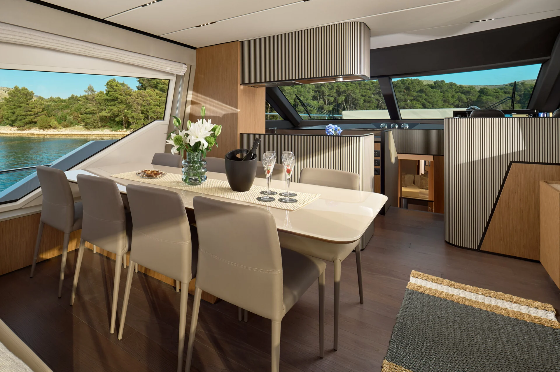 Interior of a yacht cabin featuring a dining table with six chairs, a vase of white flowers, and glasses, overlooking a view of trees and water through large windows.