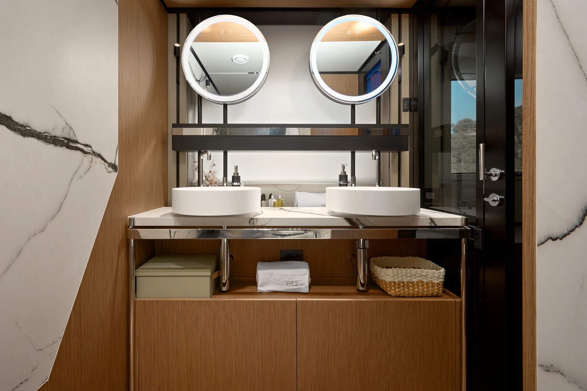 Bathroom vanity with two vessel sinks, two round backlit mirrors, and open shelving below with a storage box, a towel, and a basket.