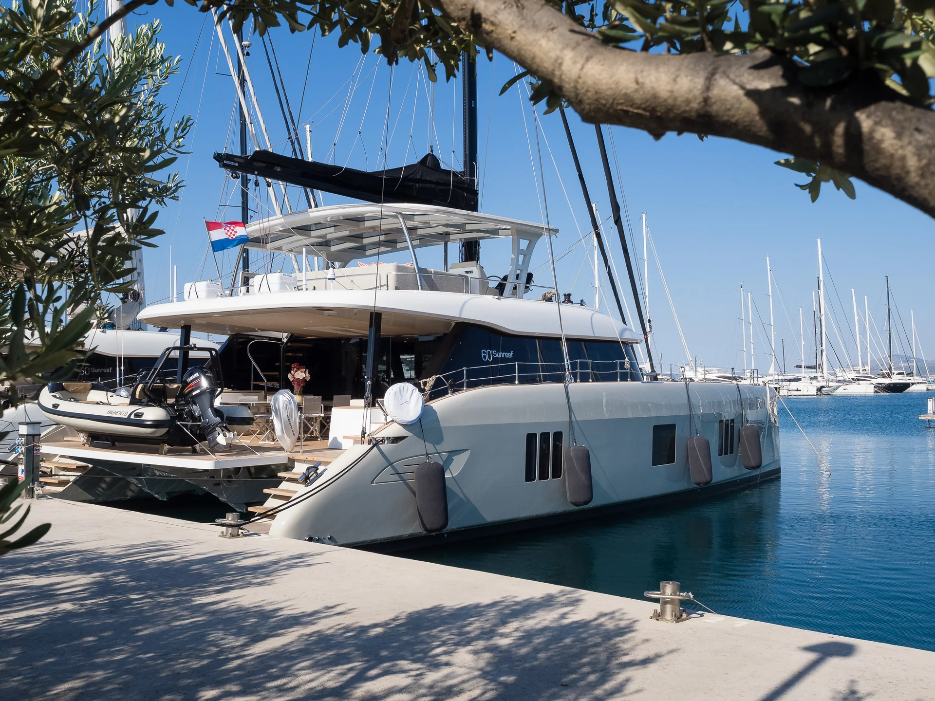 A large white catamaran yacht docked at a pier, with a small dinghy attached to the stern and a Croatian flag flying from the mast.