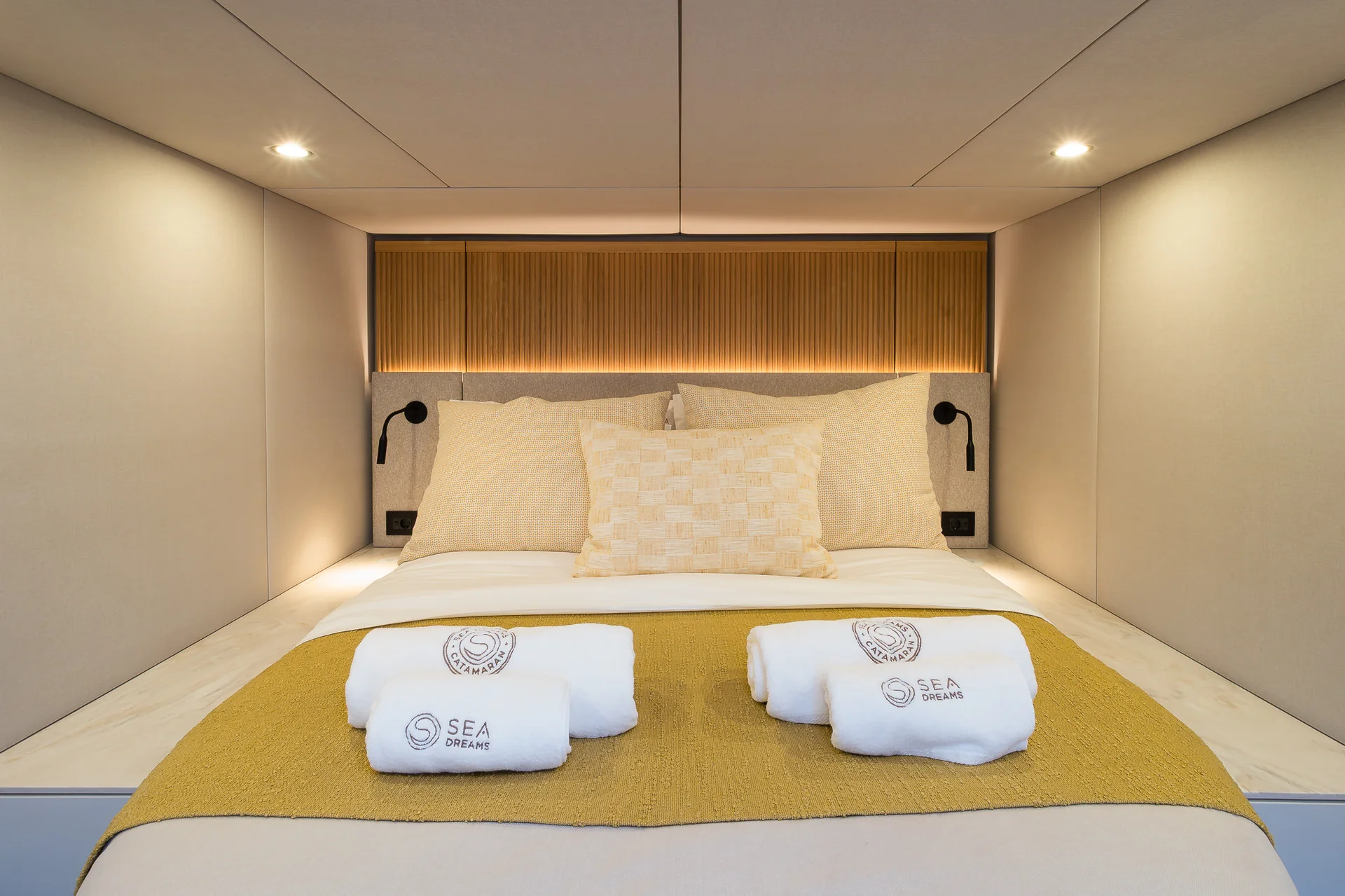 A neatly made bed with four white, branded towels folded on top of a yellow runner, situated in a minimalist bedroom cabin.