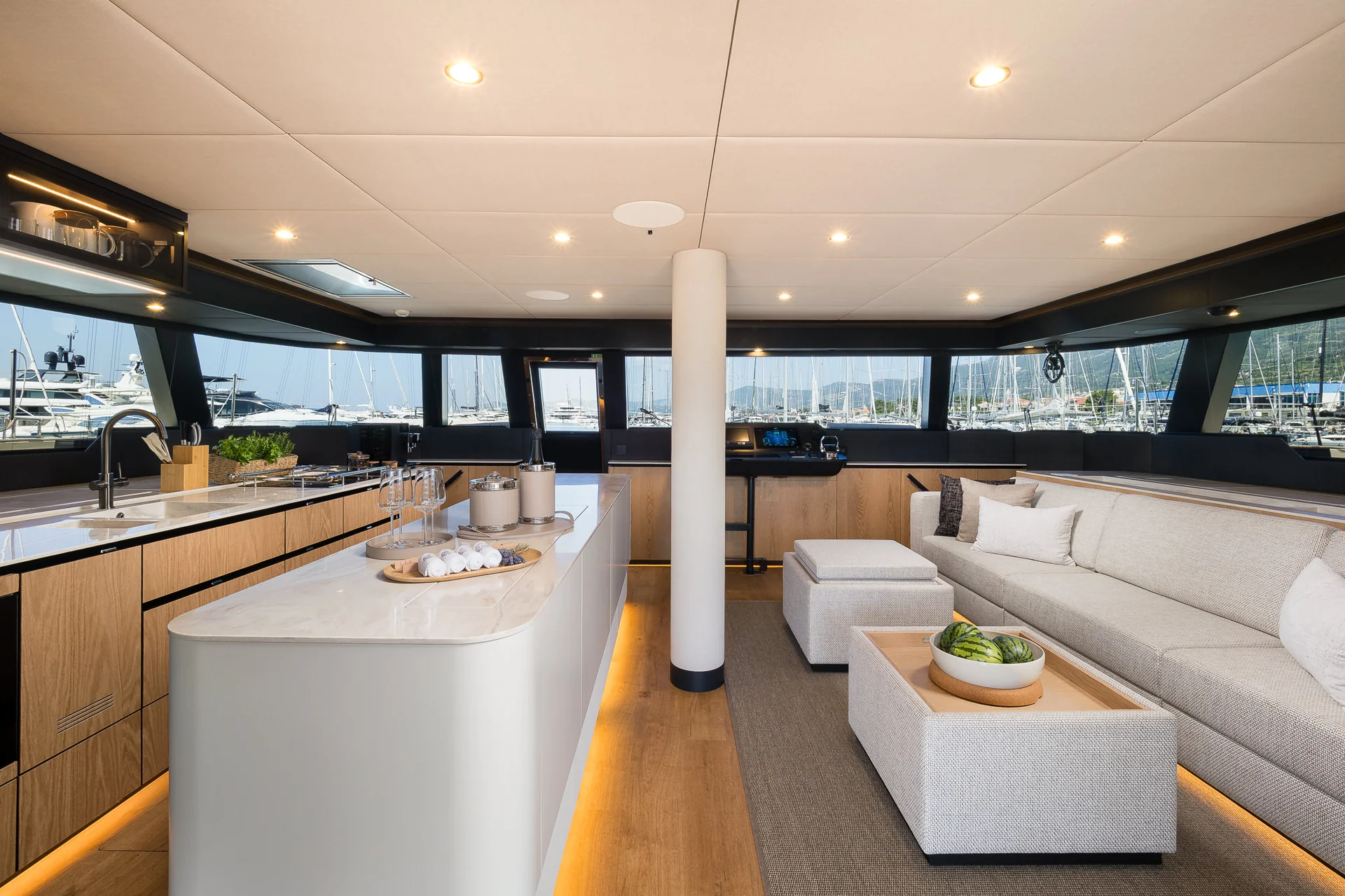 Interior view of a boat featuring a kitchen island with a white countertop and an adjacent living area with a sofa, ottomans, and views of a marina through large windows.