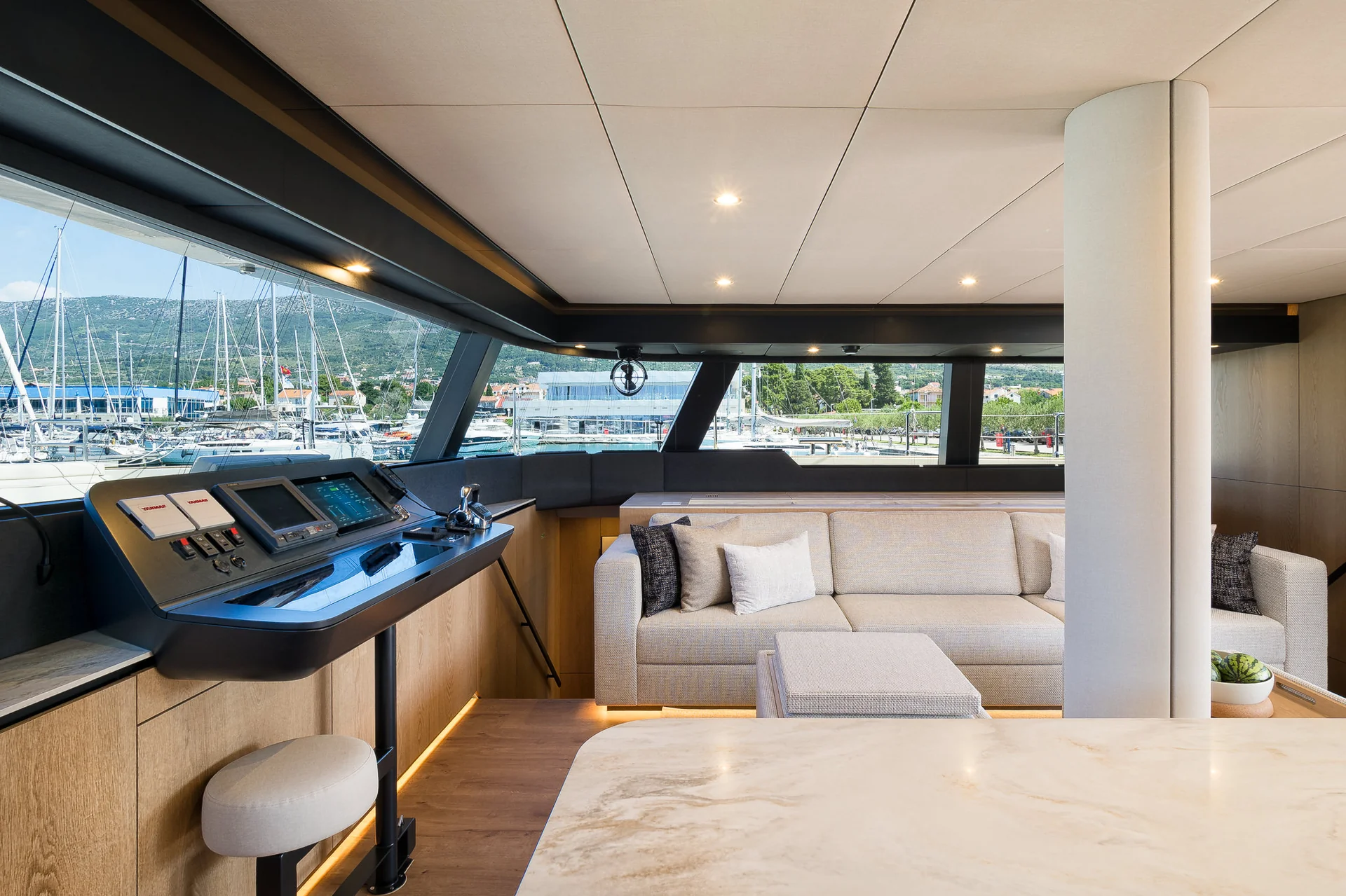 The interior of a yacht cockpit featuring a modern steering console, a plush sofa, and a large window looking out onto a marina.