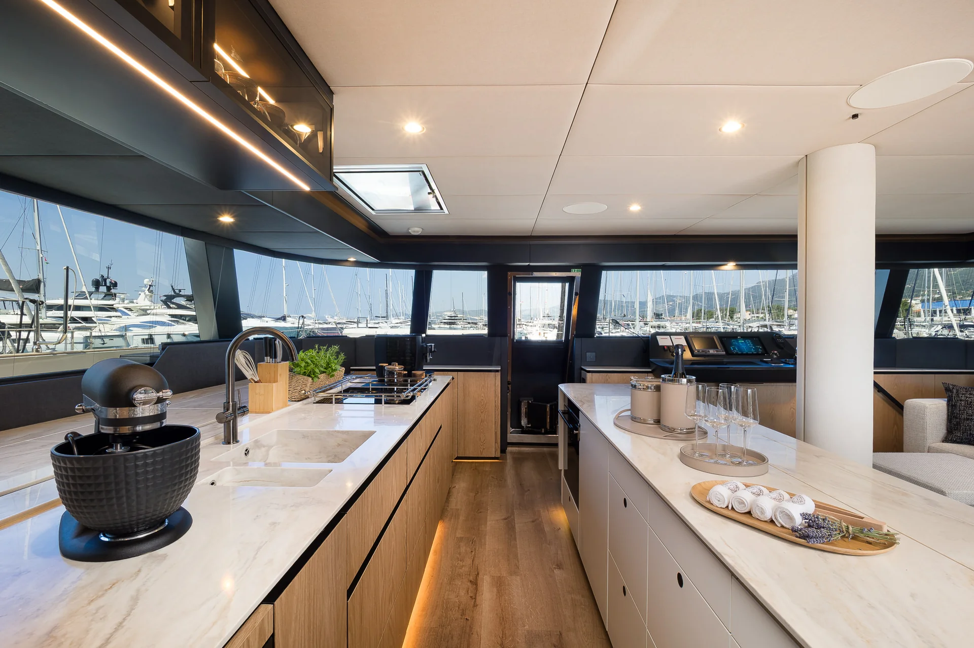 Modern kitchen inside a yacht with marble countertops, a stand mixer, and a view of a marina through large windows.